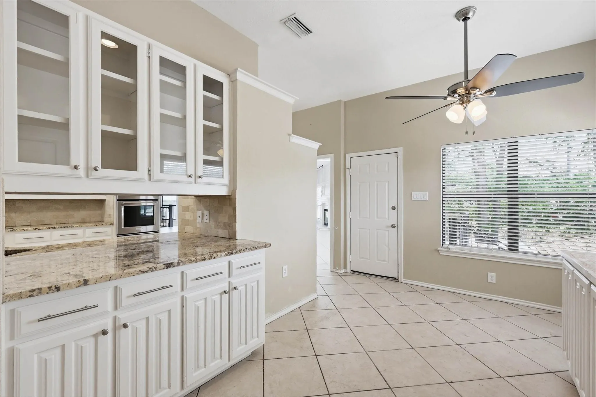White cabinetry with glass-front uppers, light-toned granite countertops, tile flooring, and a ceiling fan with integrated lighting