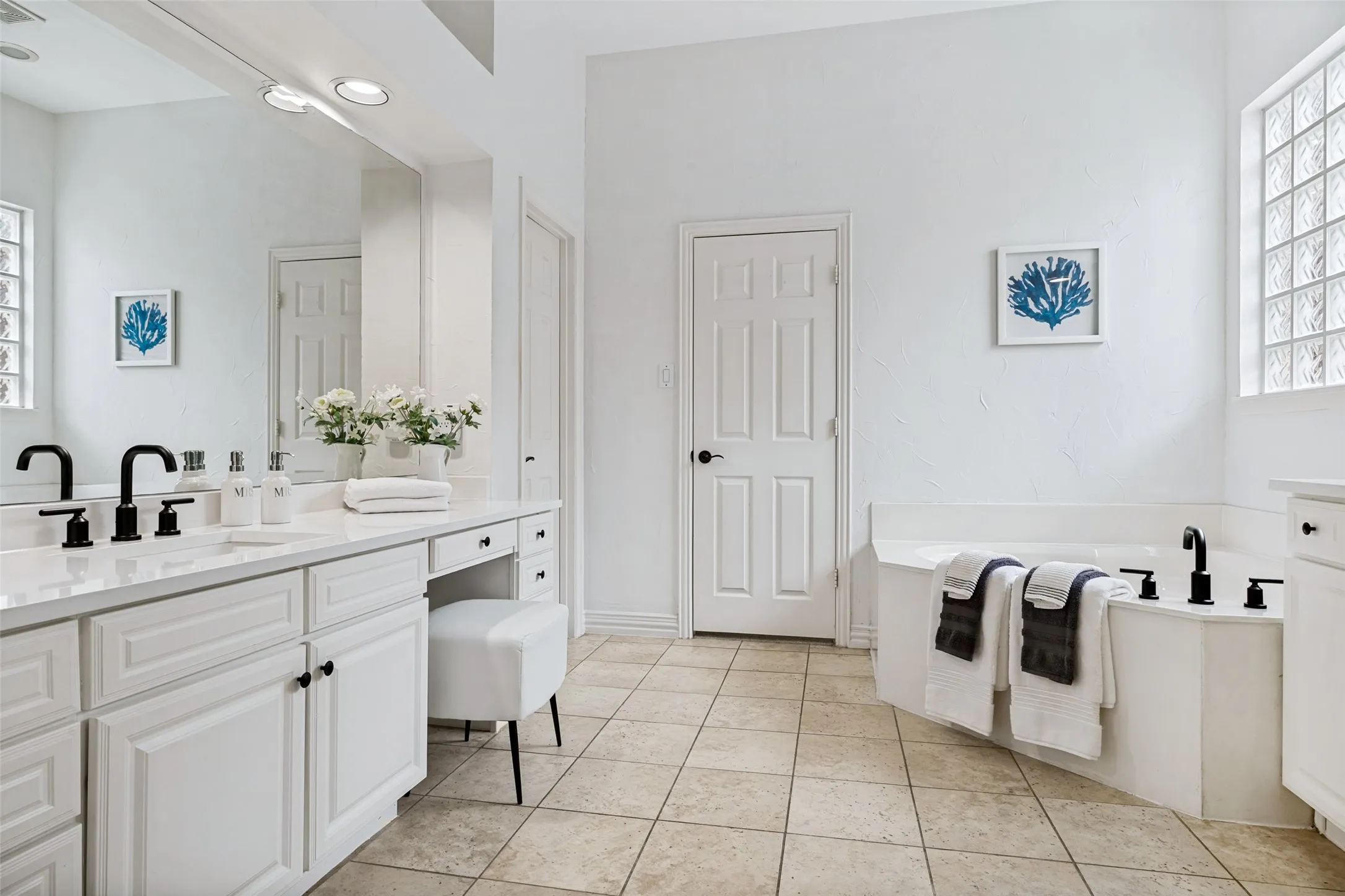 Dual vanities with white cabinetry, built-in soaking tub, tile flooring, and glass block window