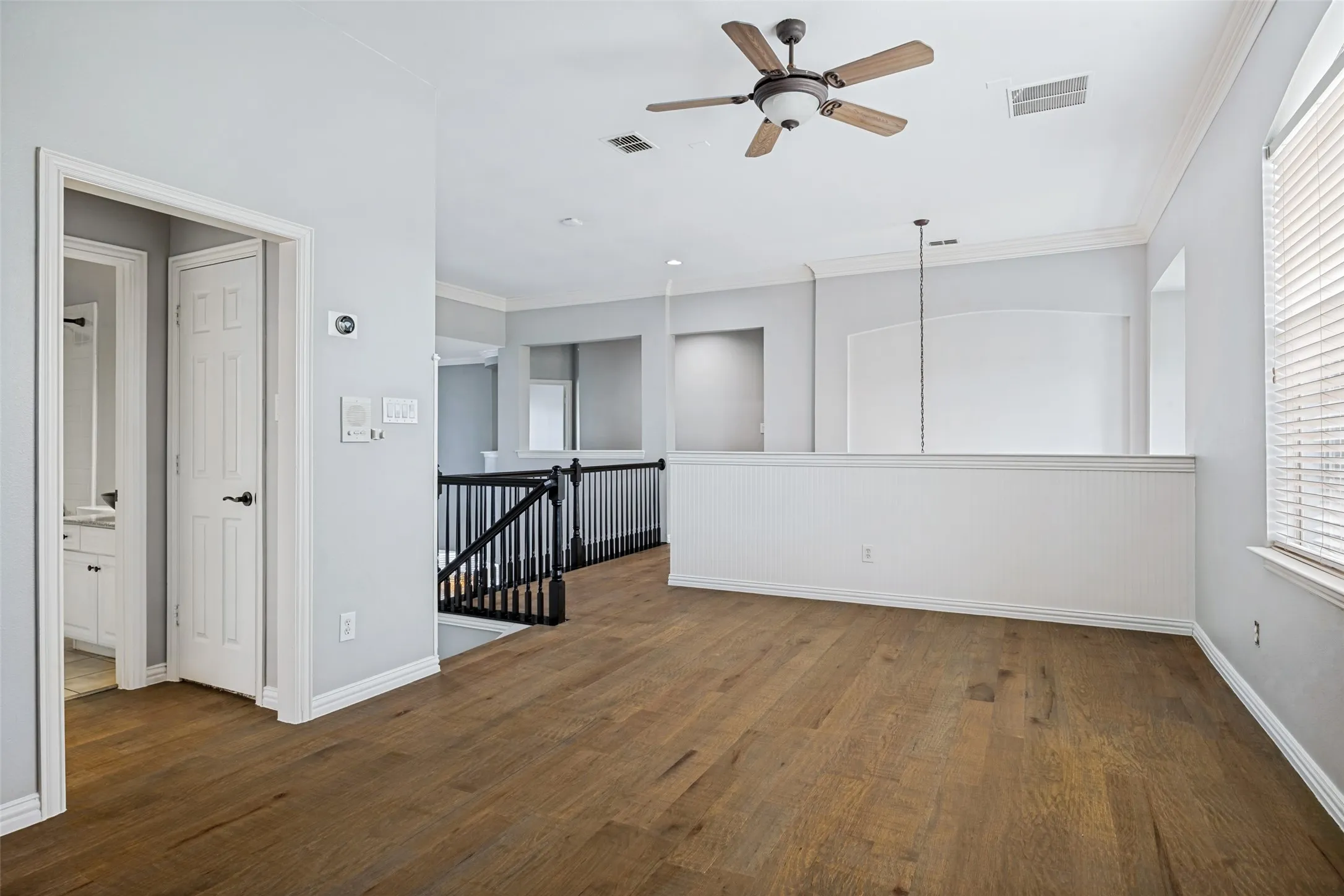 Wood-finish flooring, ceiling fan, white paneled half-wall, black metal stair railing, and a window with blinds