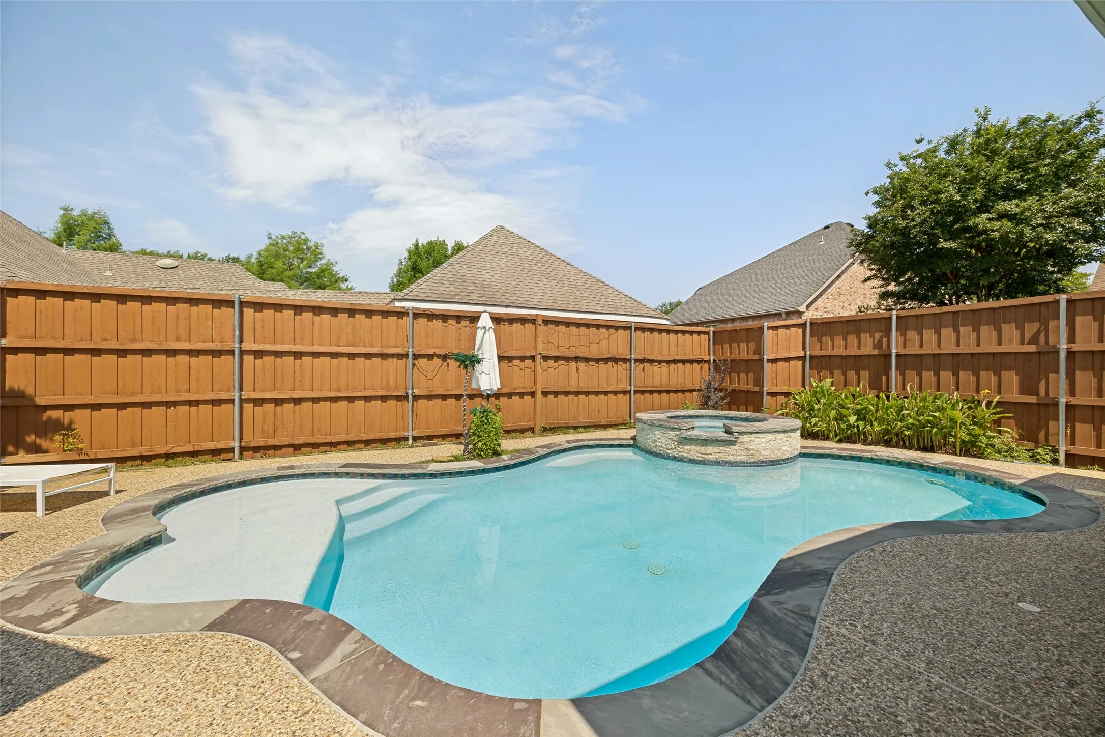 Pebble-finish pool deck, freeform swimming pool with integrated spa, and privacy fencing