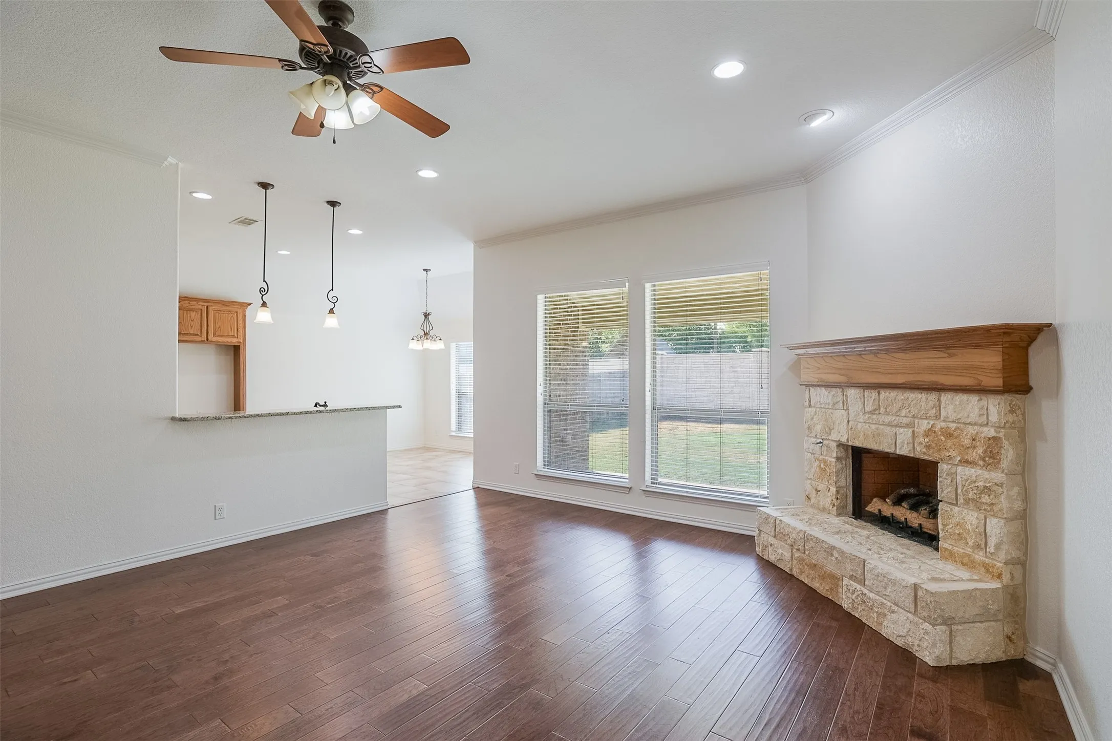 Wood-finish flooring, stone-faced fireplace with wood mantle, dual windows, ceiling fan, recessed lighting, and a breakfast bar with pendant lighting