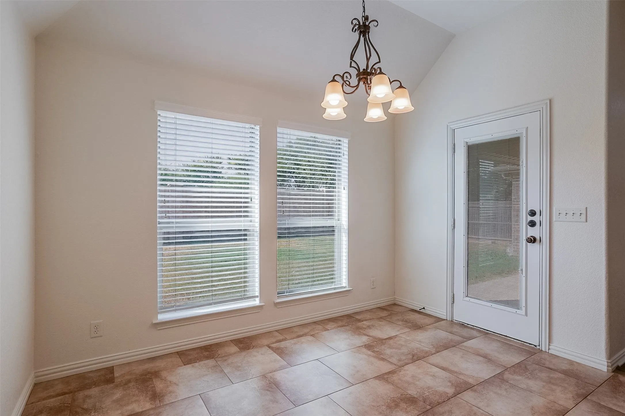 Vaulted ceiling featuring a decorative chandelier, two large windows with blinds, a glass-panel exterior door, and tile flooring