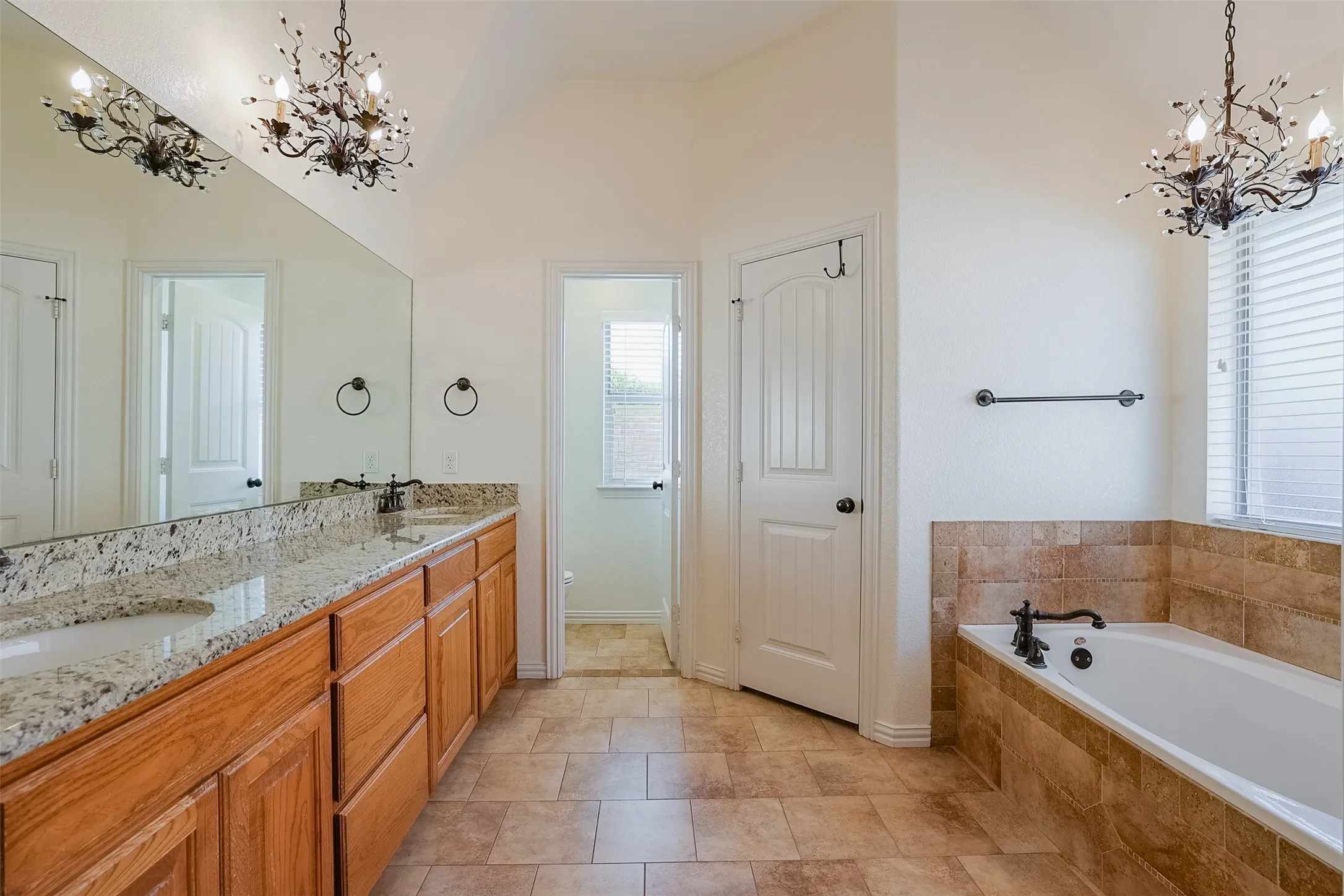 Dual vanity with stone countertops and undermount sinks, tile flooring, built-in bathtub with tile surround, and decorative chandeliers