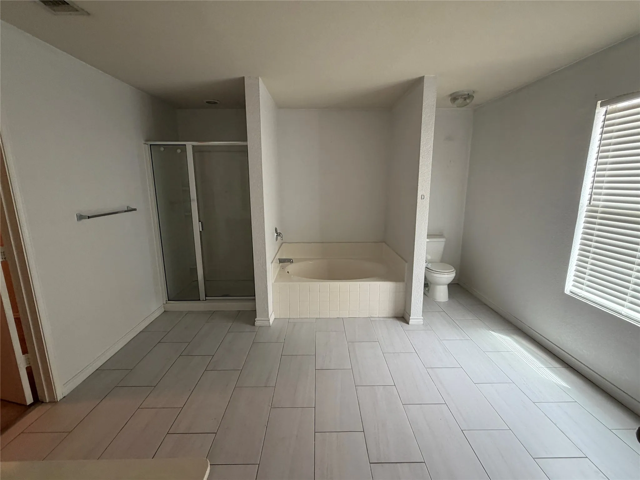 Spacious bathroom featuring light-toned plank tile flooring, a built-in bathtub, a separate shower enclosure with glass doors, and a window with blinds