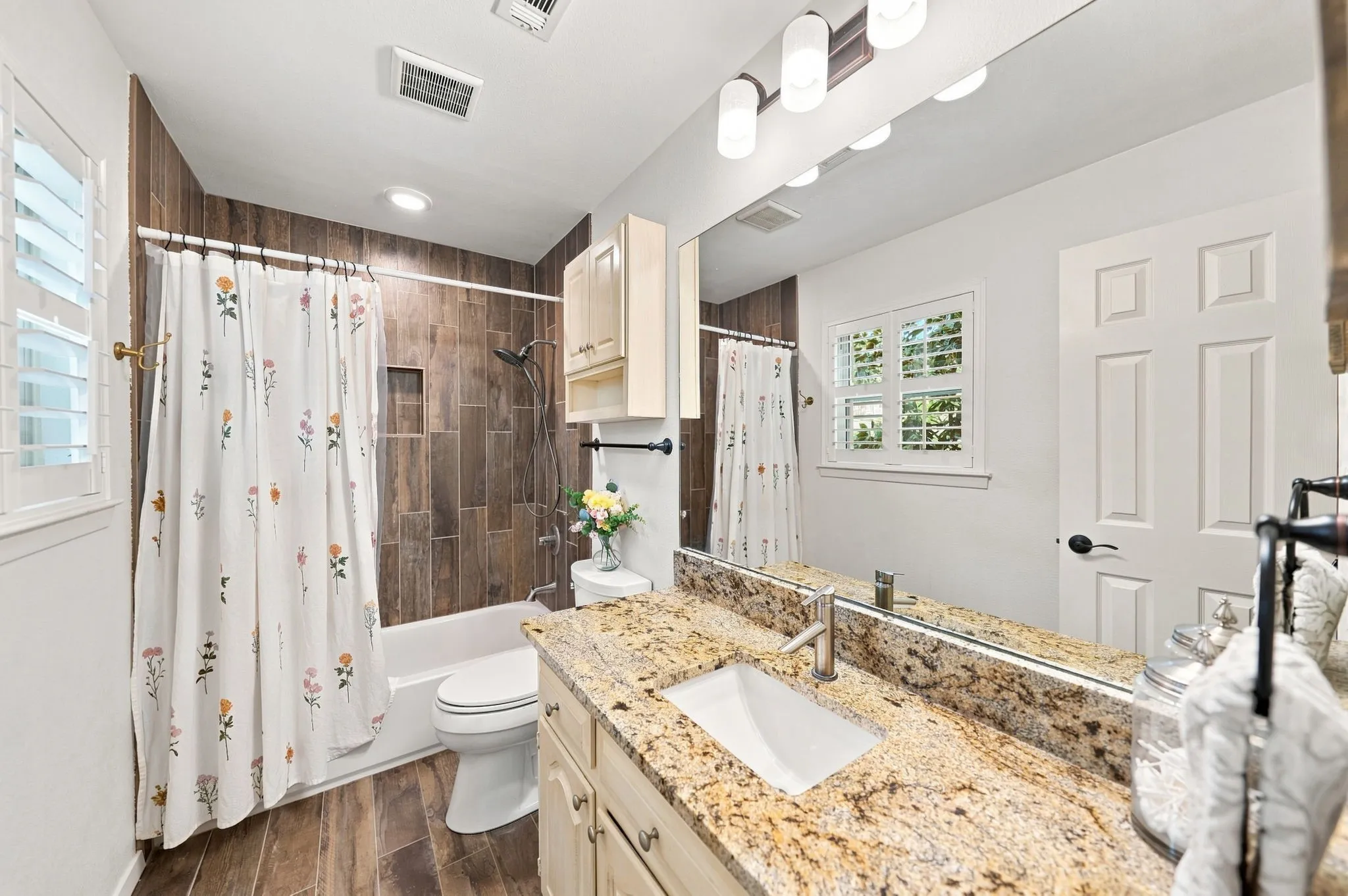 Bathroom featuring wood-finish floor tiling, a tub with a tiled shower surround and built-in niche, a vanity with a granite-style countertop, and a built-in mirror with overhead lighting