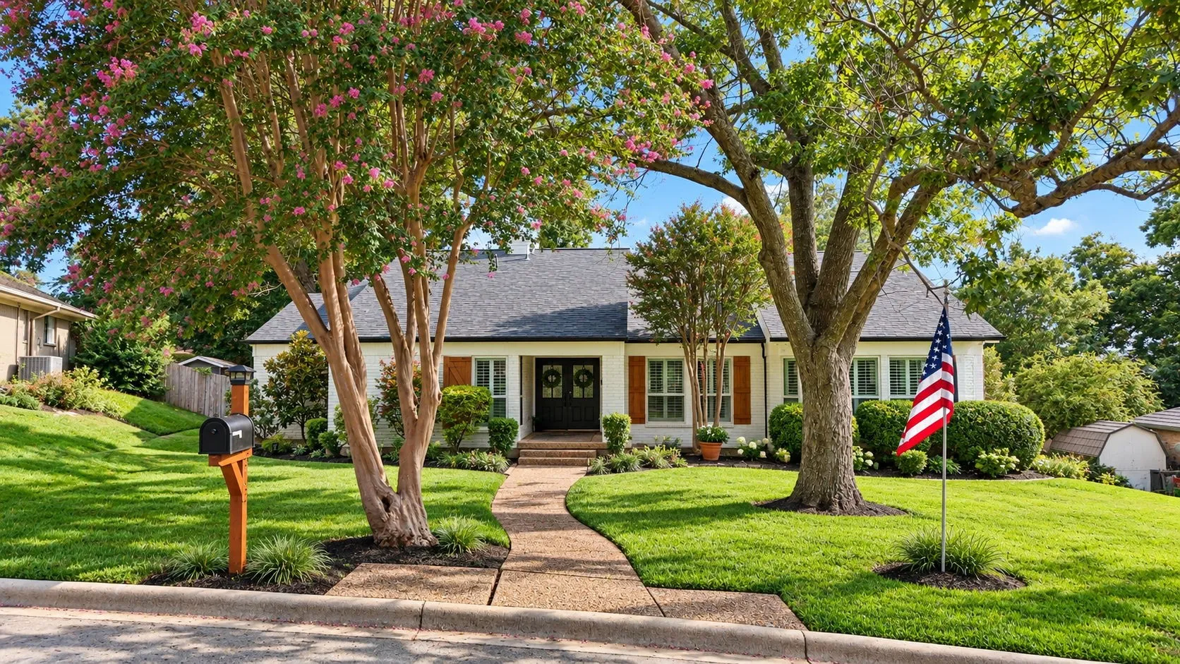 White exterior with dark shutters, dark double entry doors, a gray shingle roof, mature landscaping, and a curvilinear paved walkway