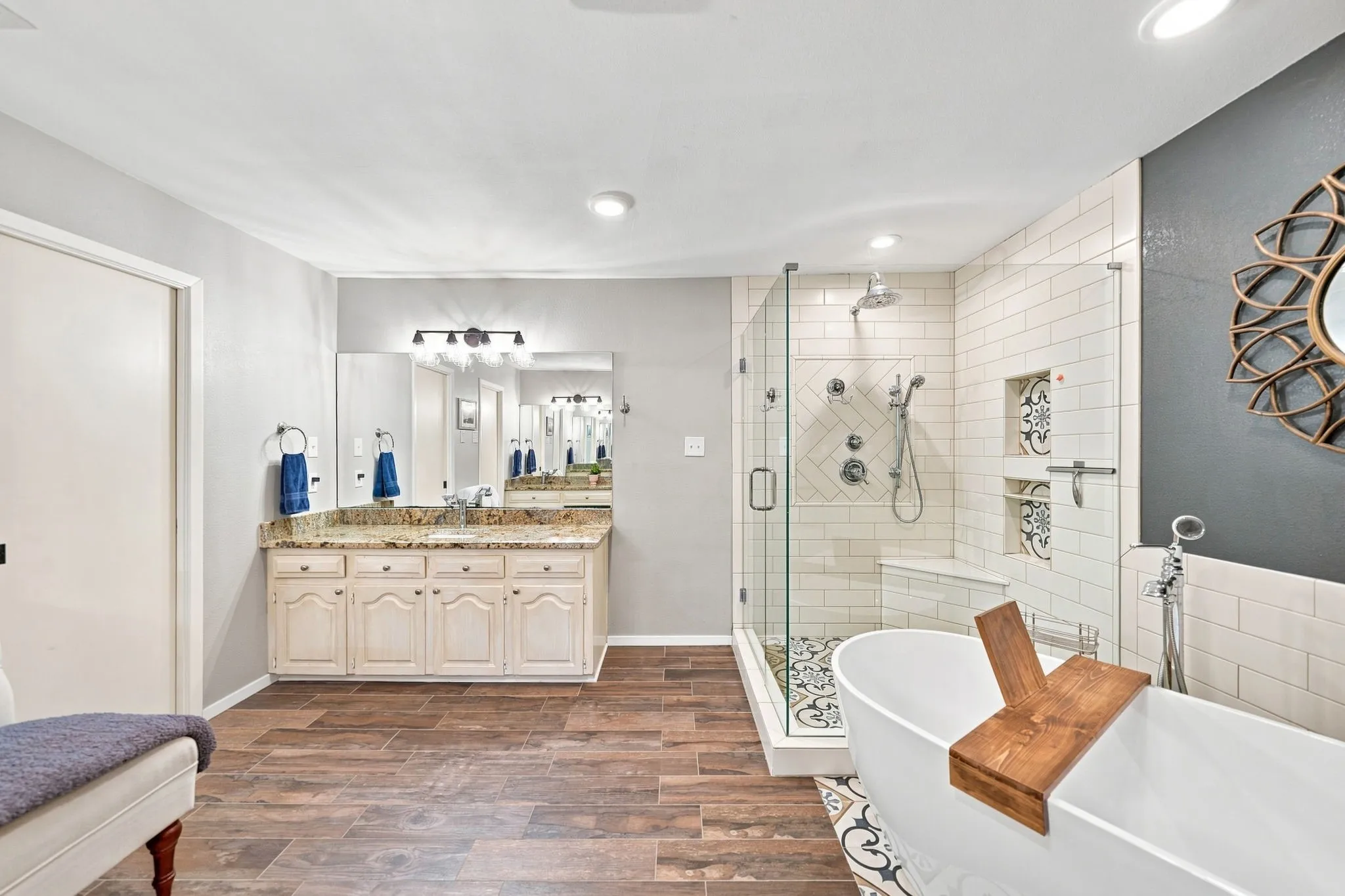 Freestanding soaking tub, walk-in shower with clear glass enclosure and subway tile surround, double vanity with granite countertop, and wood-finish flooring