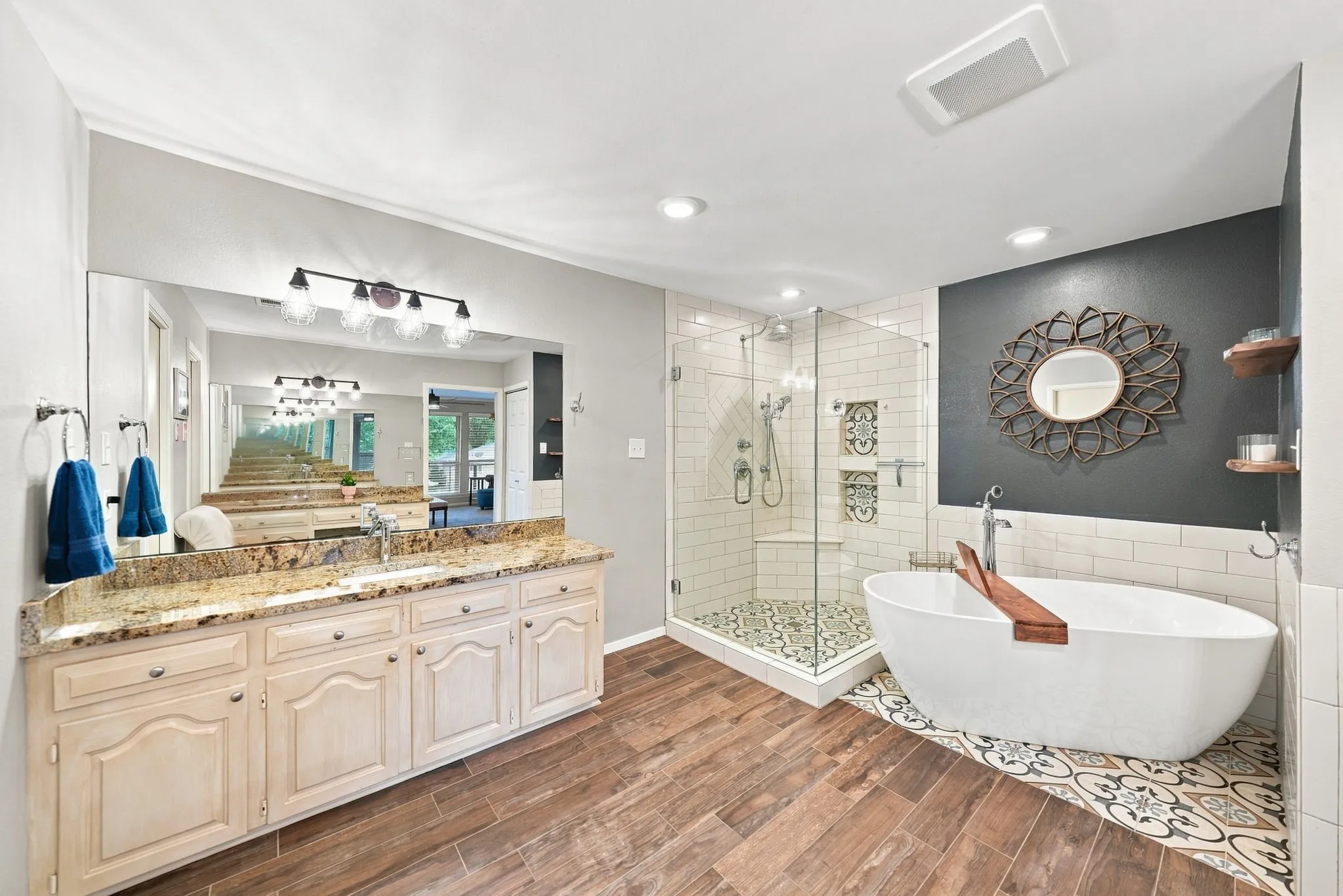 Dual vanity with stone countertop, freestanding soaking tub, glass-enclosed shower, wood-finish flooring, and decorative wall tiling