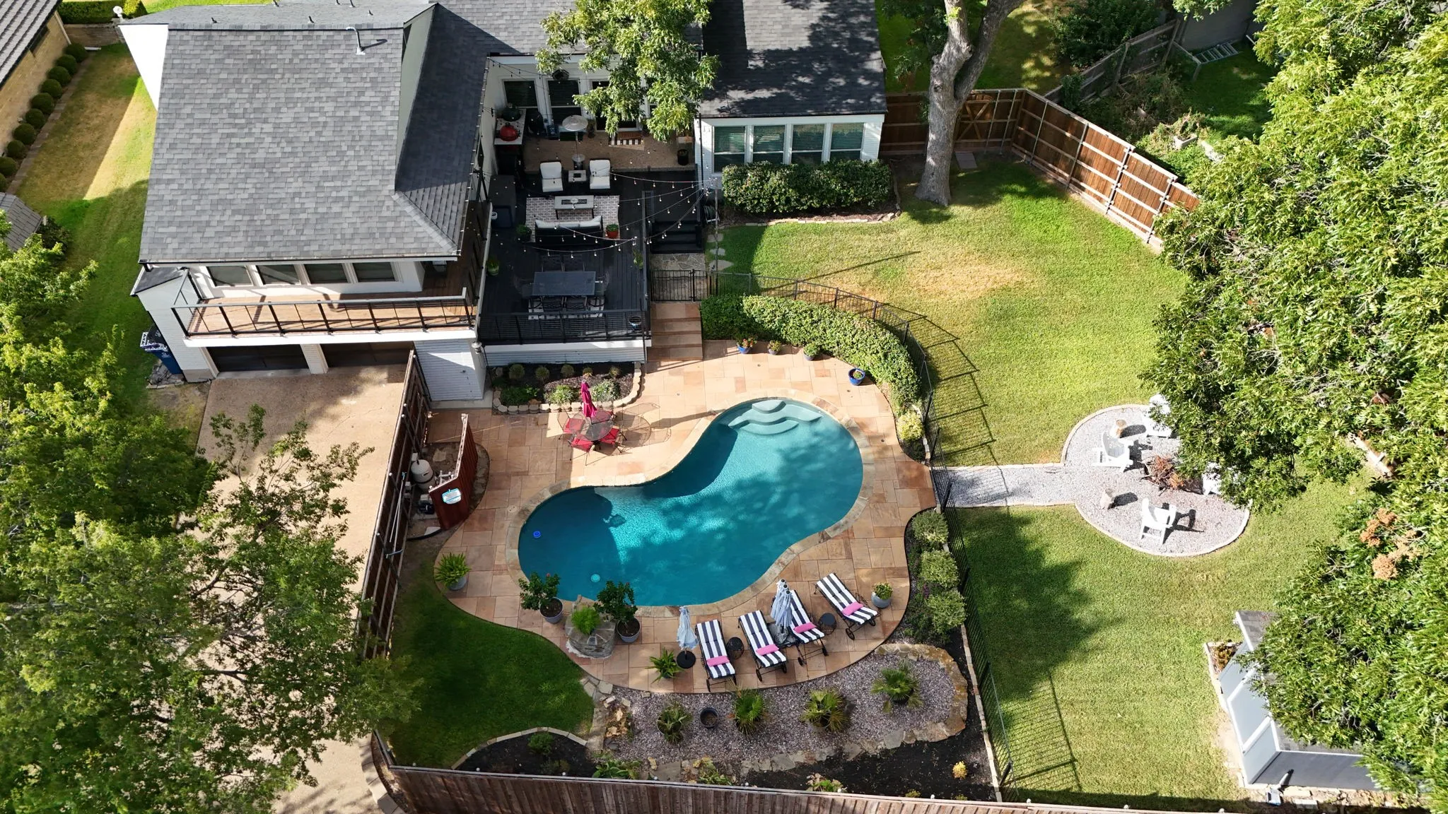 Resort-style backyard oasis featuring a custom freeform swimming pool with integrated spa, surrounding paver patio, mature landscaping with established trees, lounge seating areas, and manicured lawn space