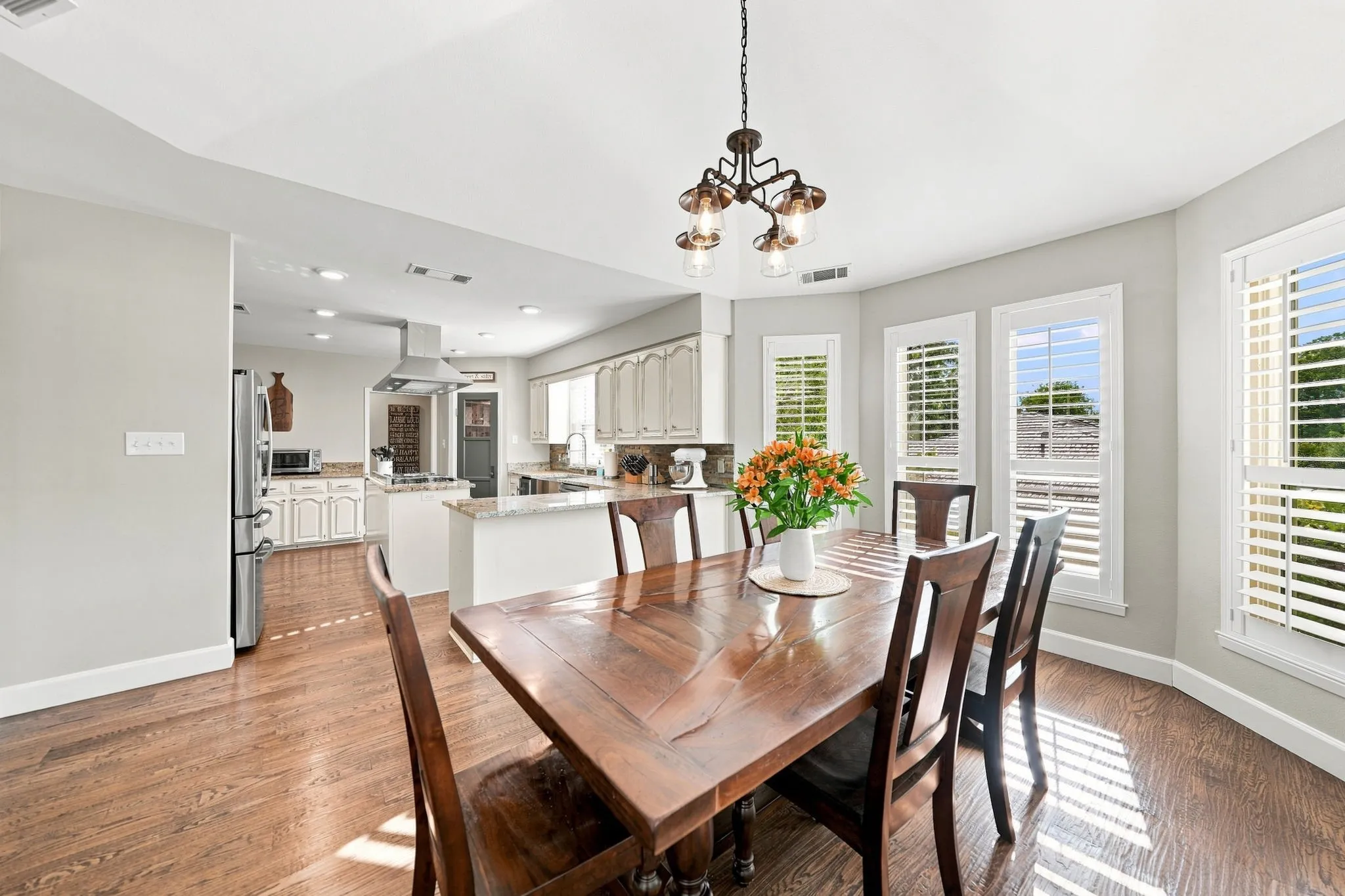 Open-concept dining area with wood-finish flooring, a decorative chandelier, and multiple windows with plantation shutters