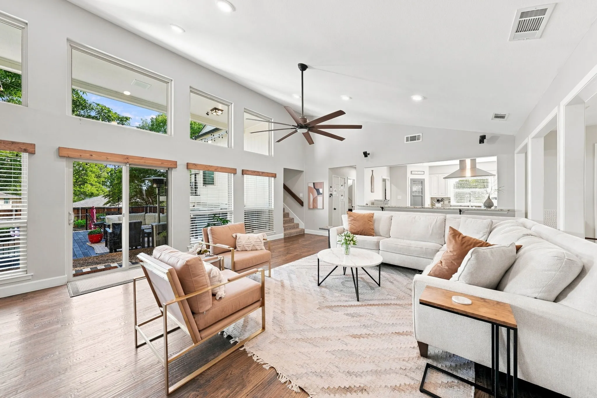 Bright, open-concept living space featuring expansive windows and glass sliding doors, hardwood flooring throughout, a ceiling fan with dark metal finish, and neutral upholstered seating arrangements