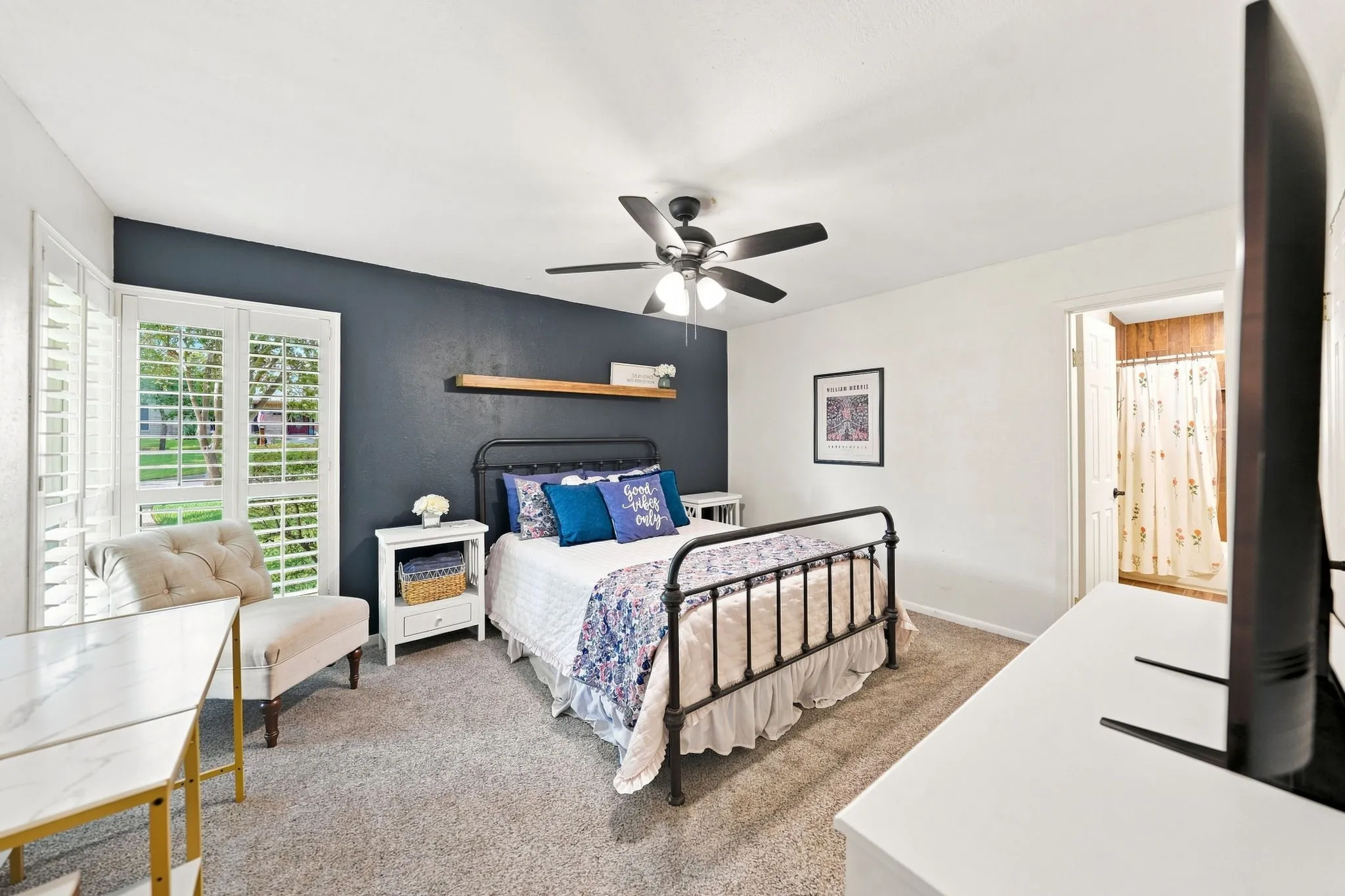 Window with interior plantation shutters, ceiling fan with integrated lighting, wood-finish floating shelf, and carpet flooring