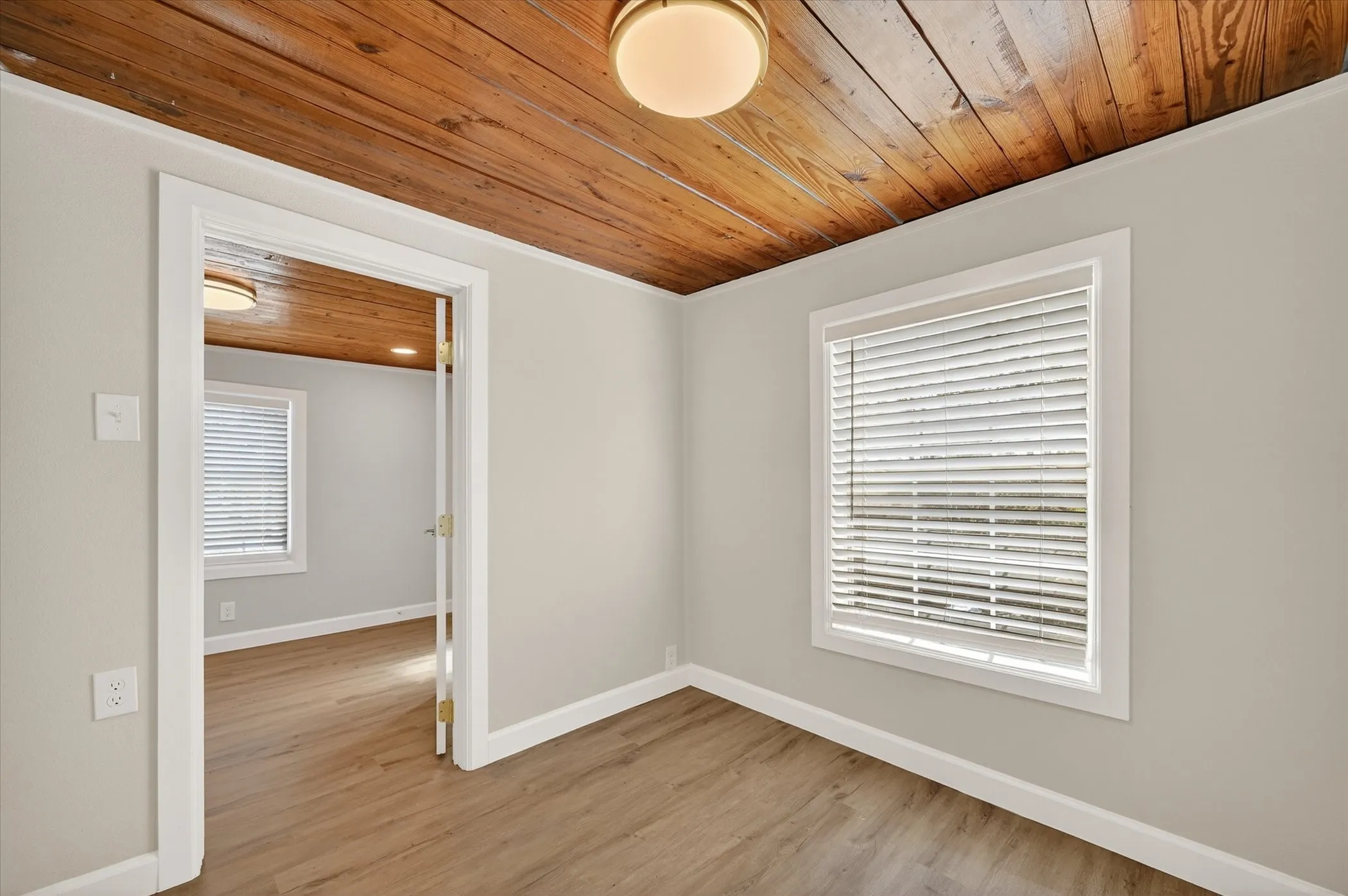 Bright corner room featuring warm wood-finish ceiling, neutral wall finishes, wood-finish flooring, and dual windows dressed with horizontal blinds allowing abundant natural light