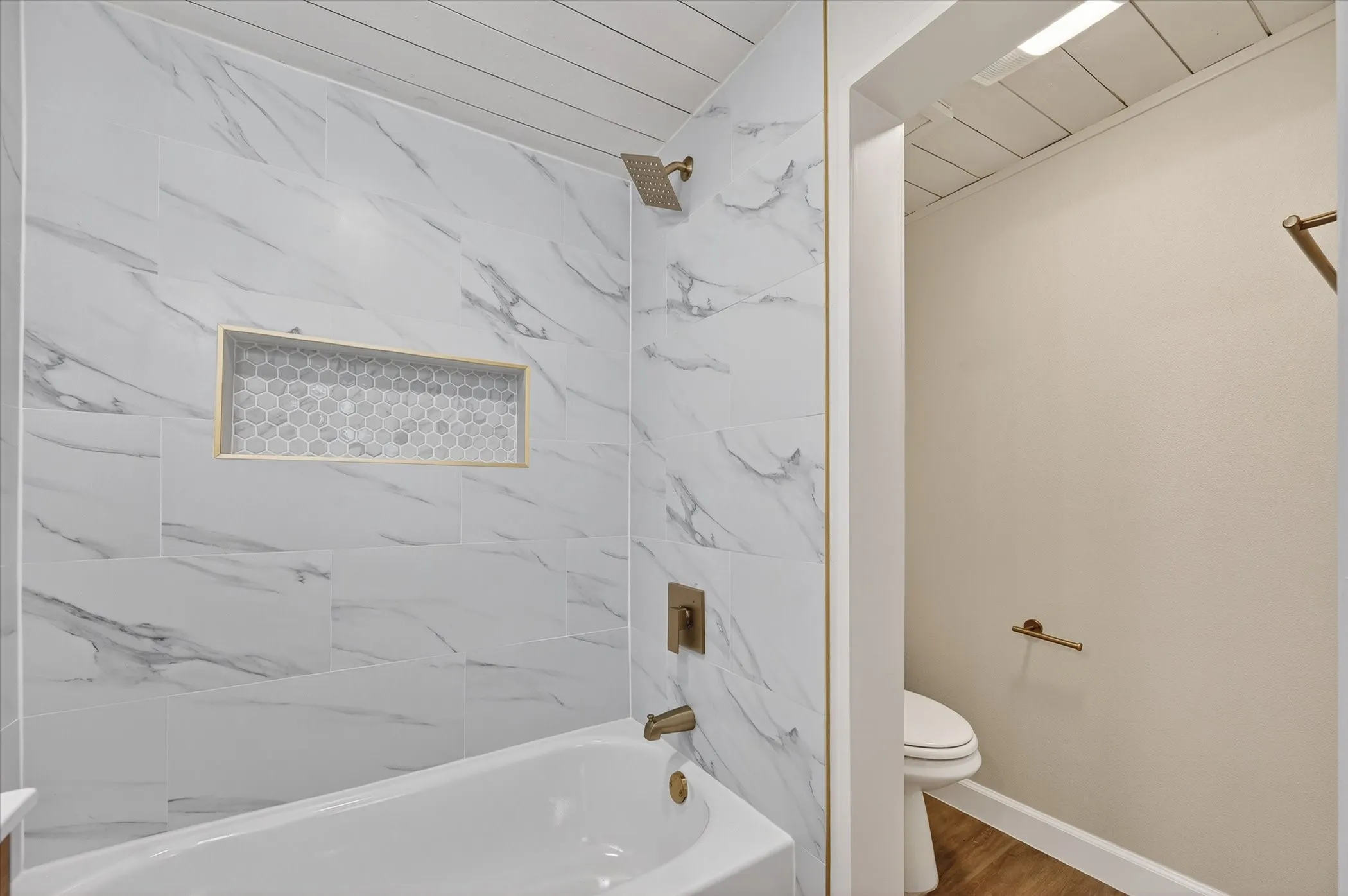Spa-inspired primary bathroom featuring a marble-look tile surround with integrated niche shelving, frameless glass enclosure, soaking tub, and brushed bronze fixtures