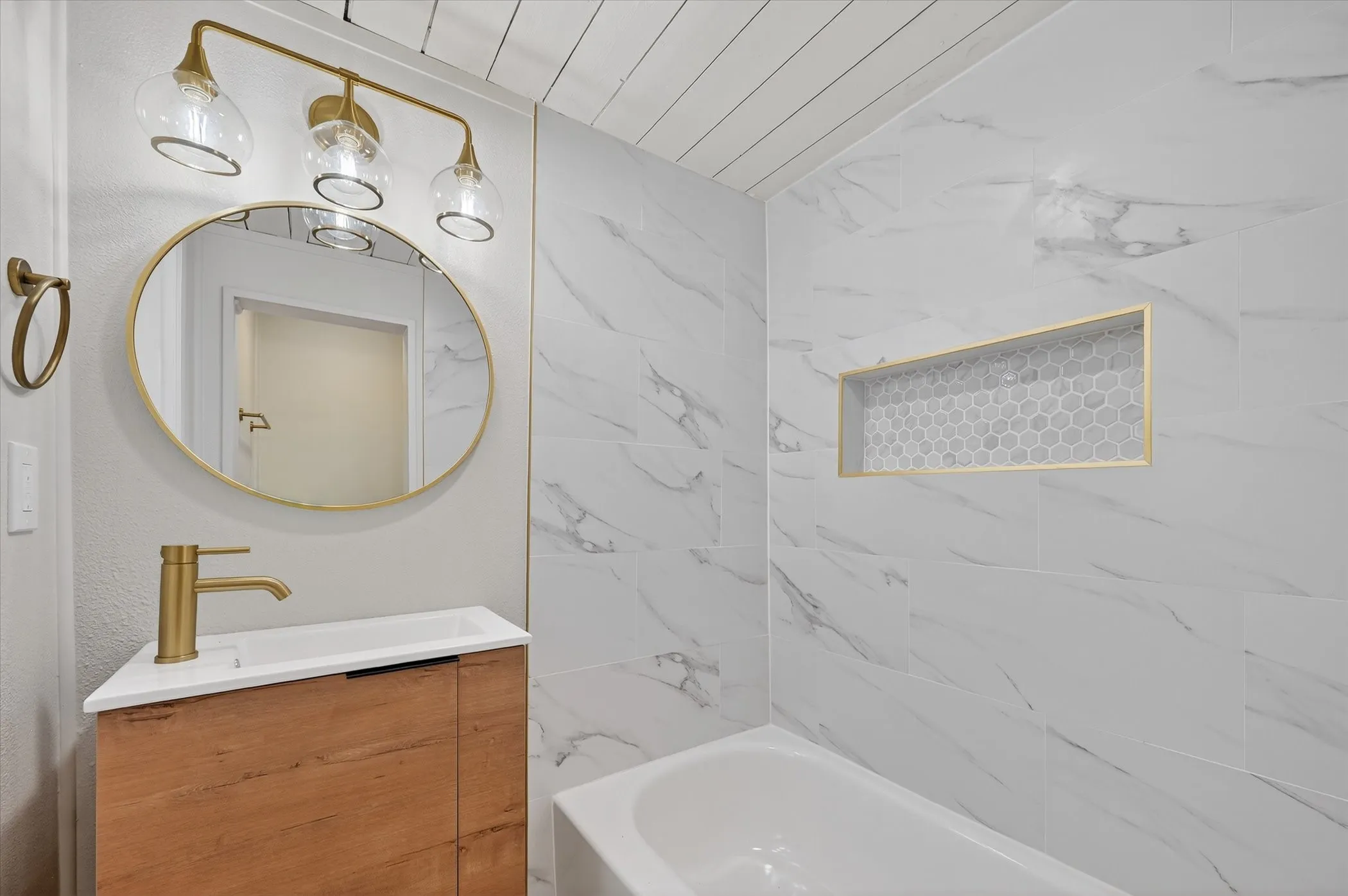 White tub with a tiled surround, integrated niche with hexagonal tile, wood-finish vanity with integrated sink, gold-finish faucet, and an oval mirror with a gold-finish frame
