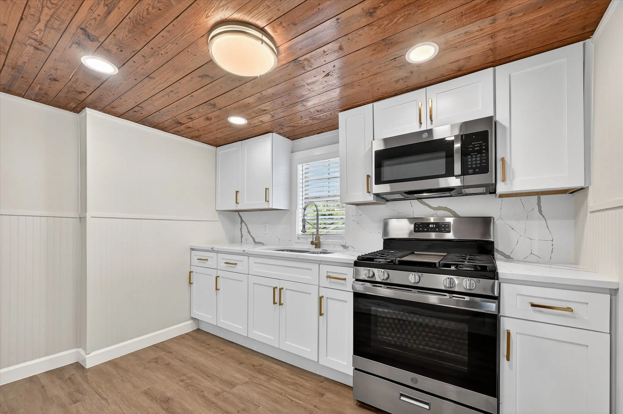 Modern kitchen featuring white shaker-style cabinetry with brushed gold hardware, stainless steel gas range and matching microwave, quartz countertops with marble-look veining, recessed and flush-mount ceiling lighting, and warm wood-finish ceiling detail throughout