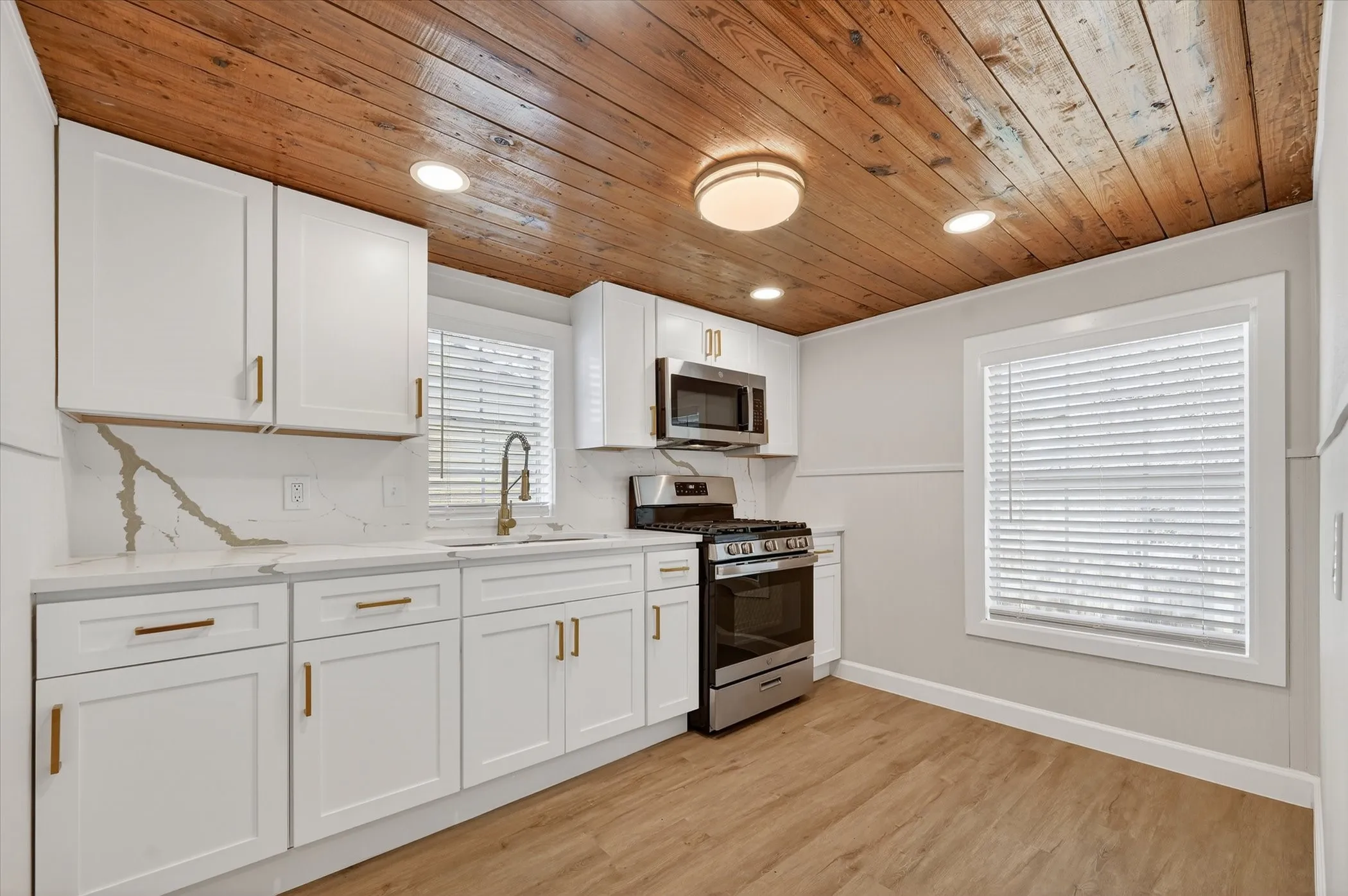 Modern kitchen featuring white cabinetry with brushed gold hardware, quartz countertop with marble-look backsplash, stainless steel gas range and built-in microwave, wood-finish plank ceiling with recessed lighting, and wood-look flooring throughout