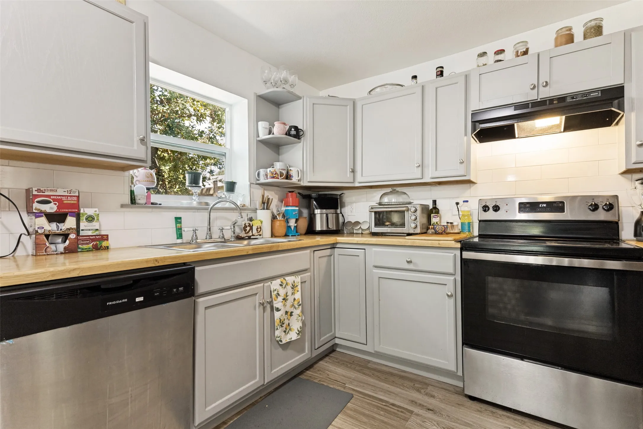 Light gray cabinetry with brushed nickel hardware, stainless steel appliances, wood-finish countertops, white subway tile backsplash, and light wood-look flooring