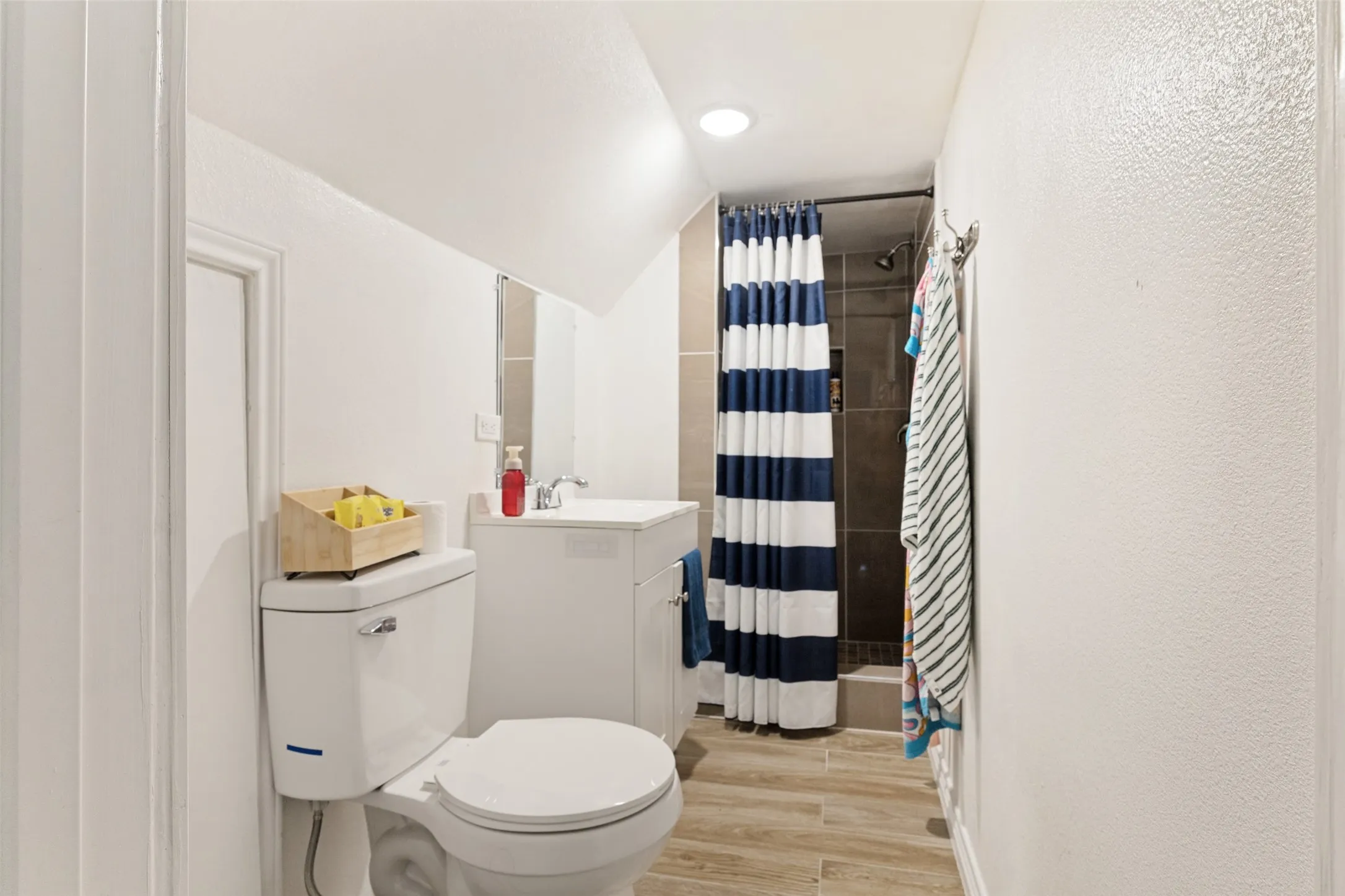Bathroom featuring a white toilet, white vanity with integrated sink, wood-look flooring, and a shower with dark tiled walls