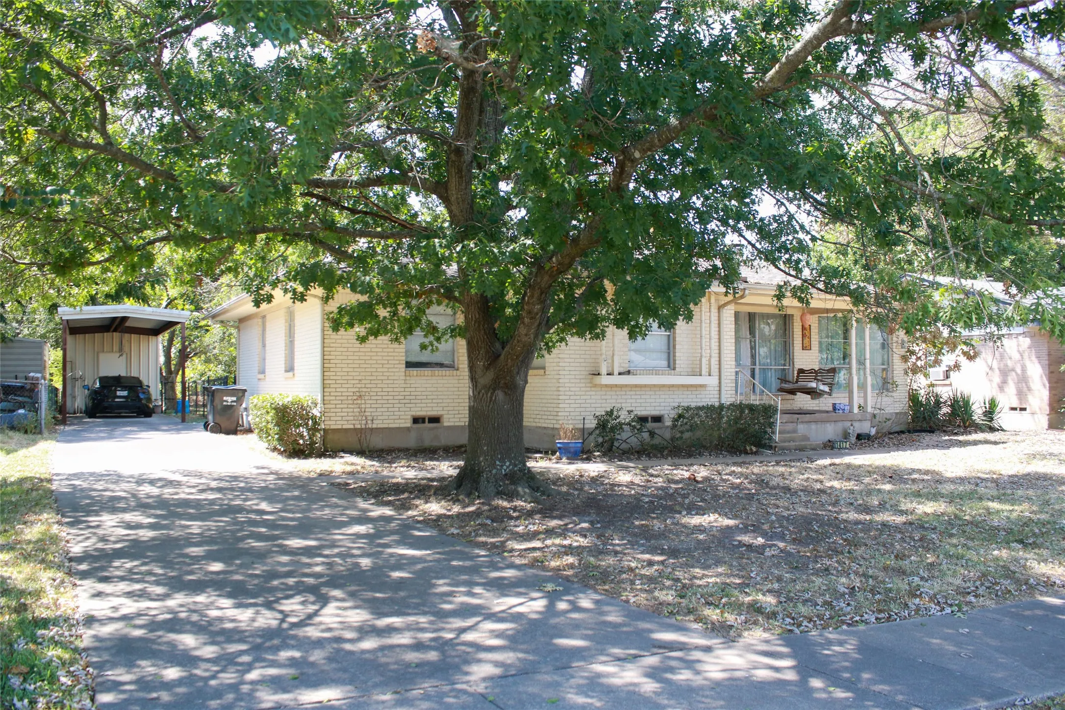 Cream brick exterior, mature trees, concrete driveway, covered carport, and covered front porch
