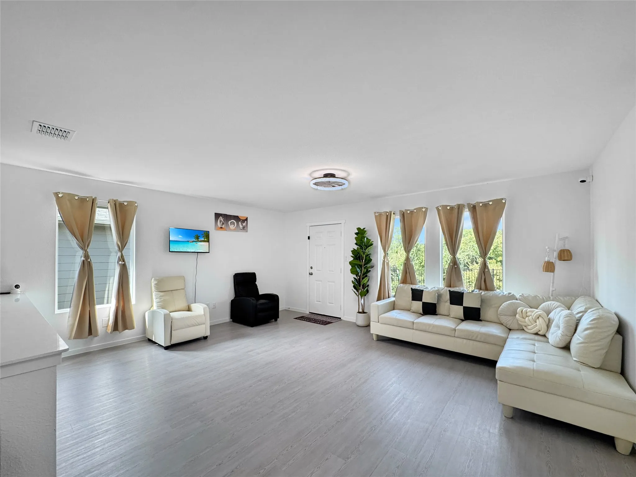 Spacious open-concept living area with light wood-finish flooring throughout, multiple sets of glass sliding doors with decorative curtain panels providing natural light, cream-toned upholstered seating, wall-mounted flat-screen television, recessed ceiling lighting, and abundant windows framing outdoor greenery