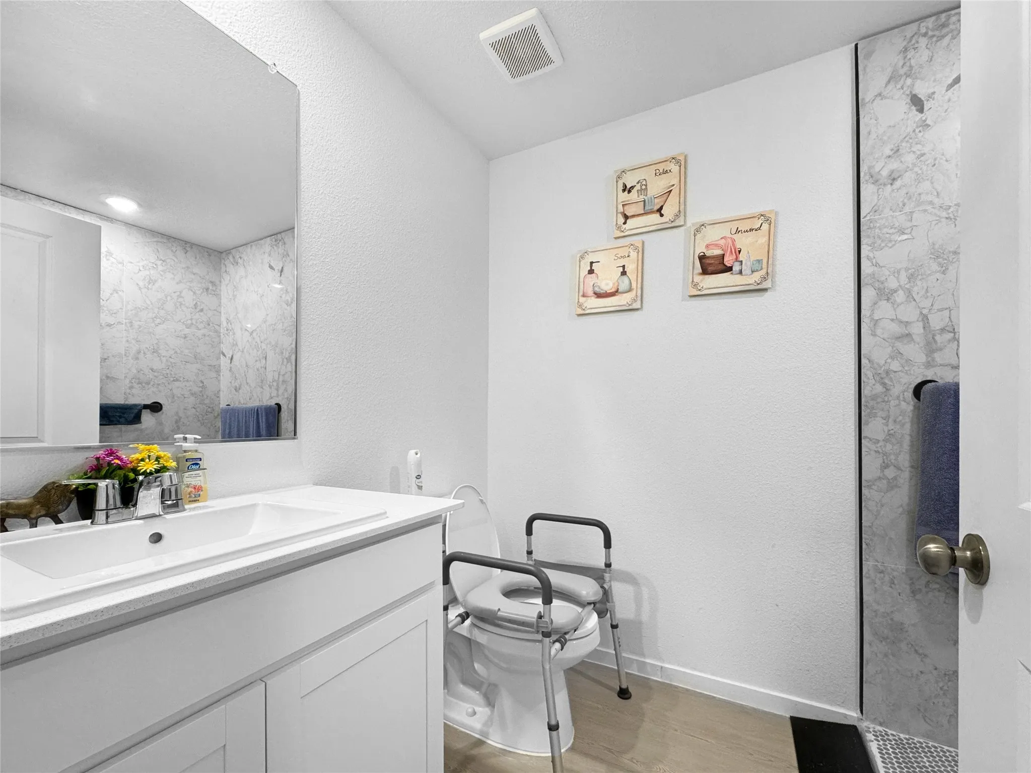 Modern bathroom featuring a white vanity with integrated sink and ample countertop space, large framed mirror, polished chrome hardware, light wood-finish flooring, and a walk-in shower with stone-look tile surround and glass enclosure