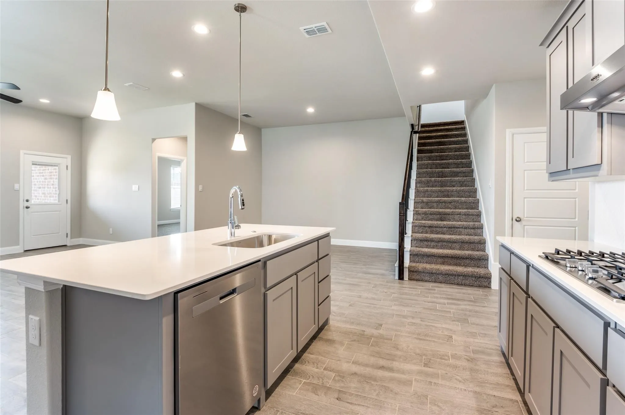 Kitchen island with sink and integrated dishwasher, recessed lighting, wood-finish flooring, full staircase with carpeted treads, and gray cabinetry