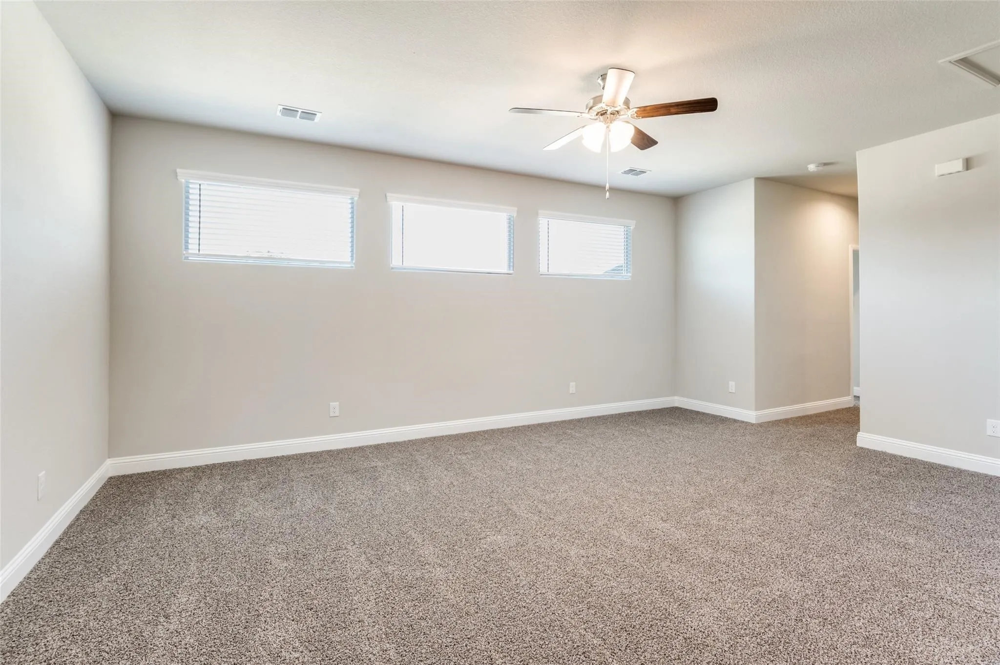 Spacious room featuring neutral carpeting, three horizontal windows with blinds, a ceiling fan with wood-finish blades, and recessed lighting