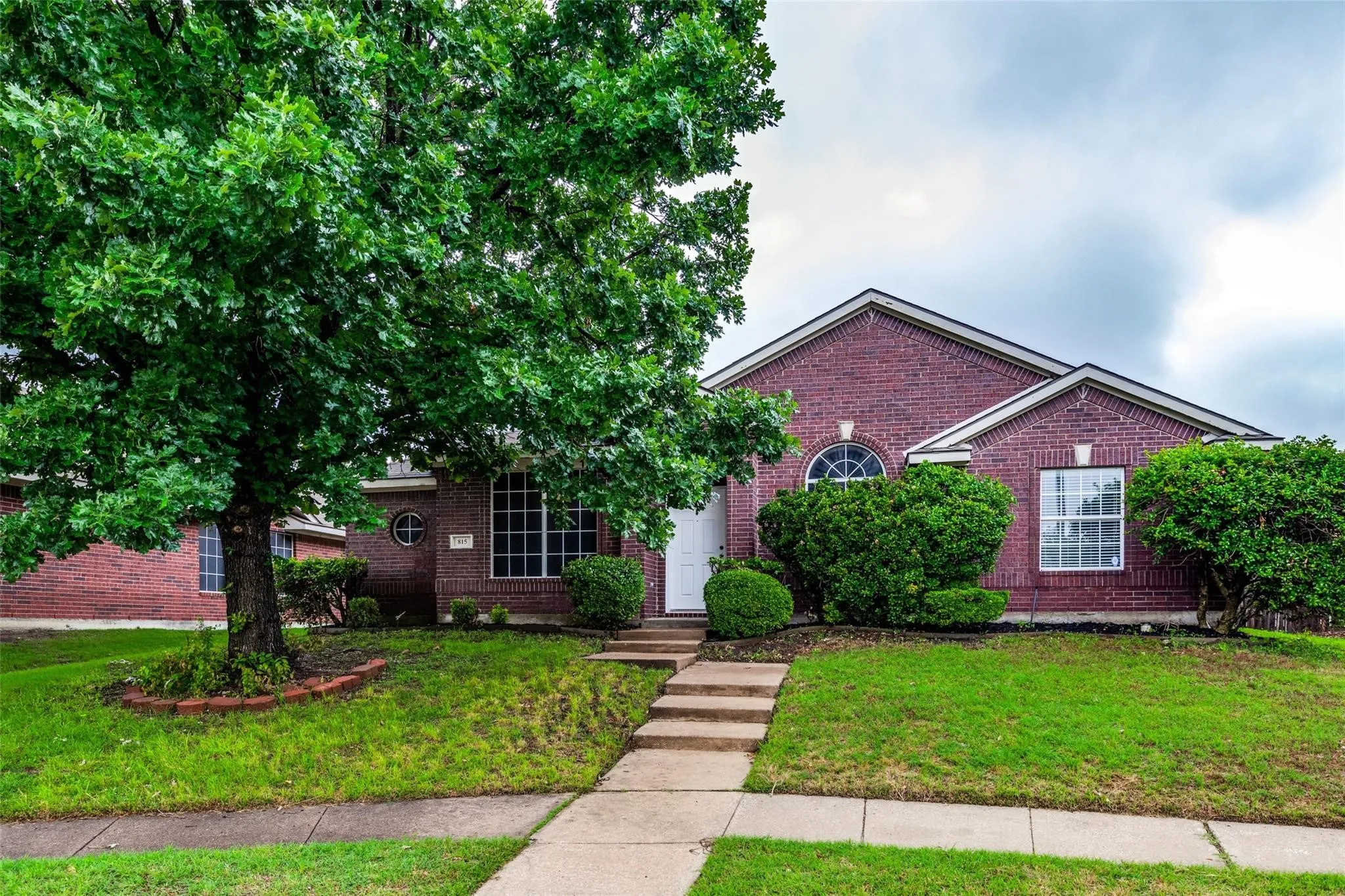 Brick exterior featuring a gabled roofline, mature tree, concrete walkway, and landscaped front yard with shrubs
