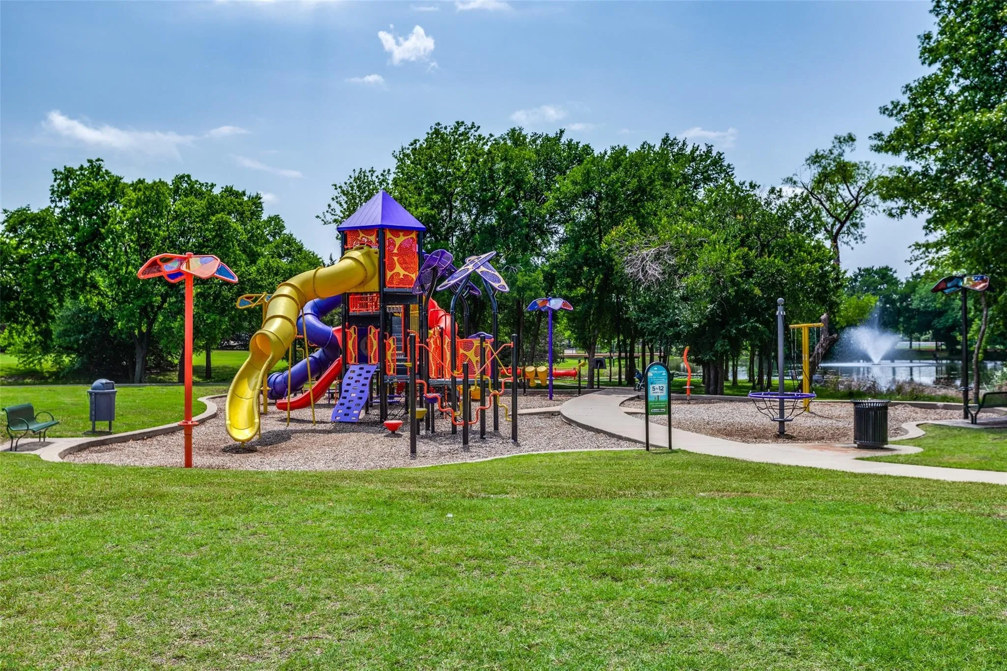 Expansive park area featuring a colorful playground with slides and climbing structures, surrounded by mature trees