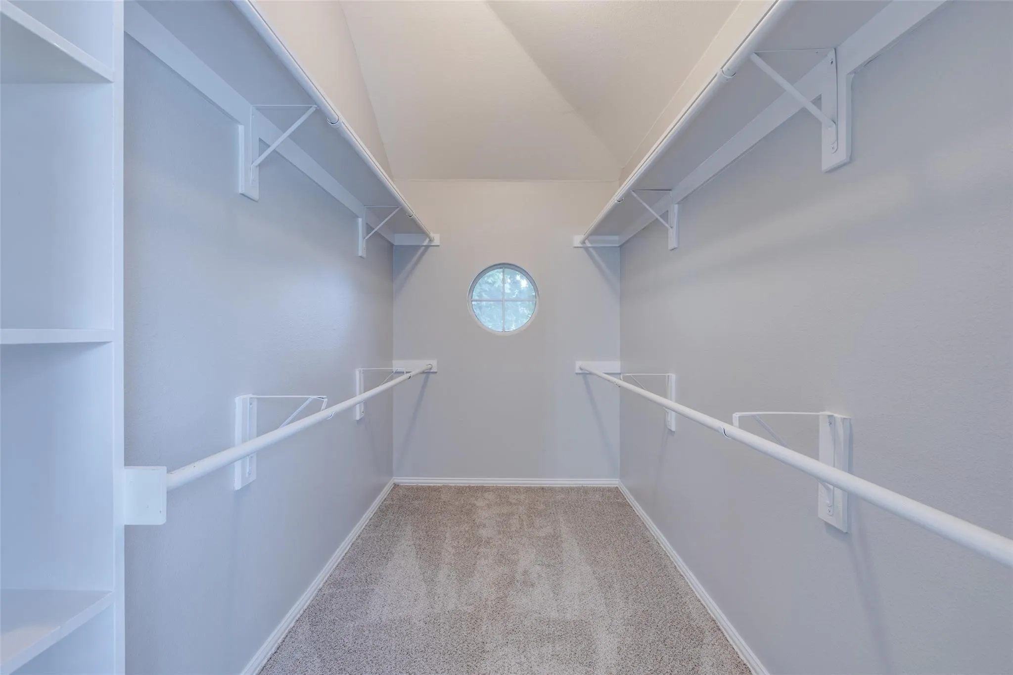 Walk-in closet featuring built-in shelving, dual hanging rods, upper storage shelves, a circular window, and light-toned carpeting