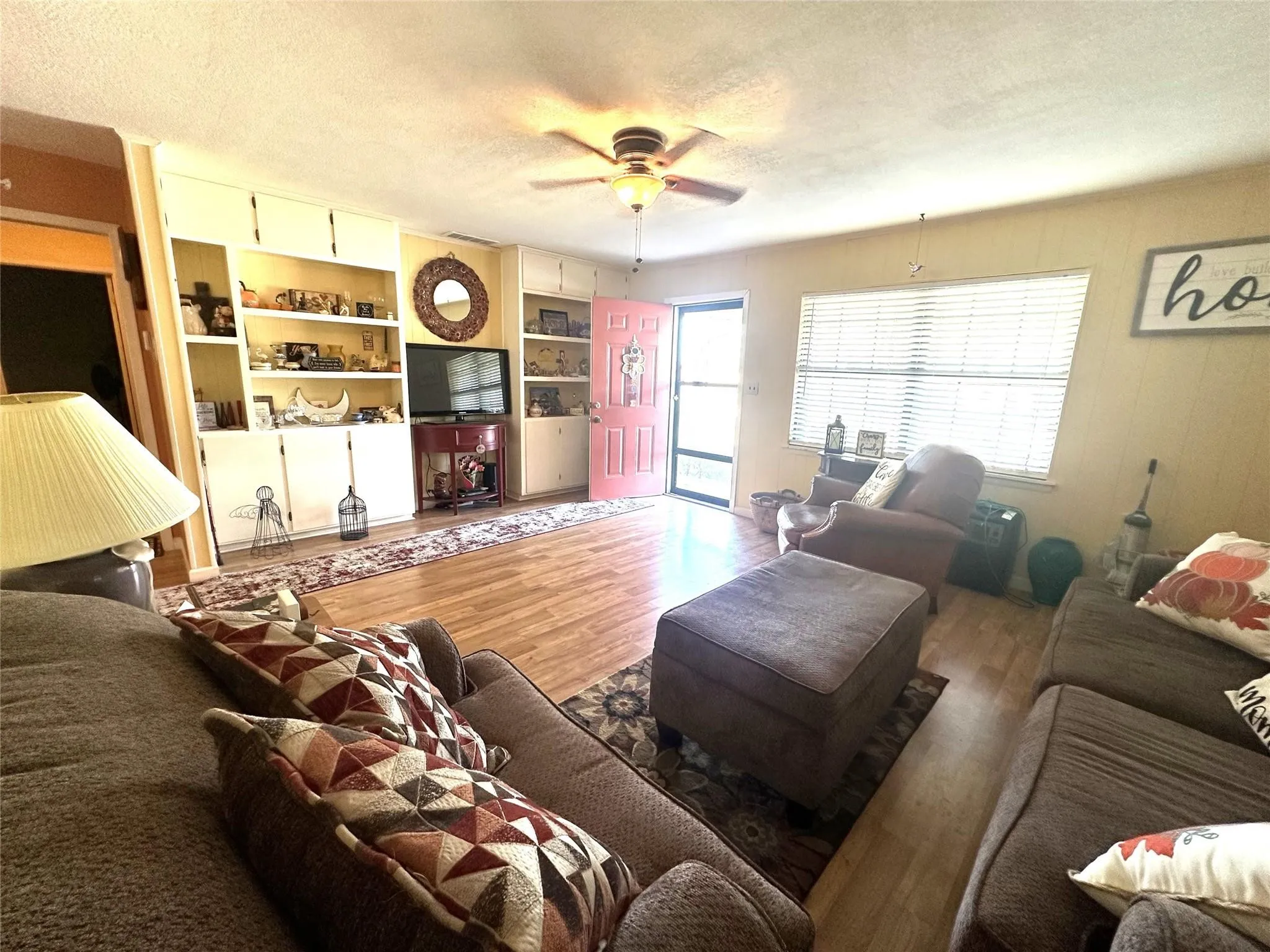 Living area featuring wood-finish flooring, built-in shelving, a ceiling fan, and large windows with blinds