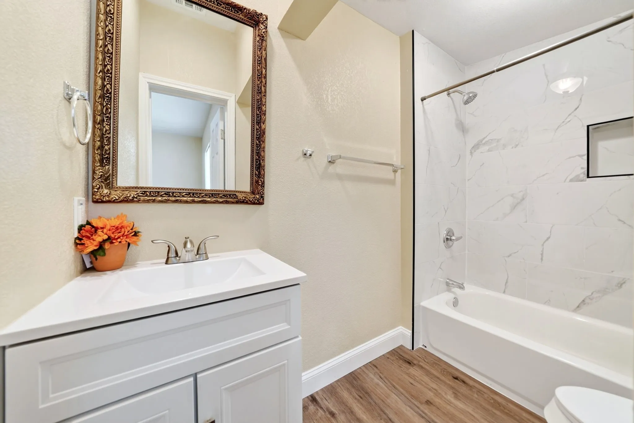 White vanity with an integrated sink and chrome faucet, wood-finish flooring, and a shower-over-tub combination with large format tile surround