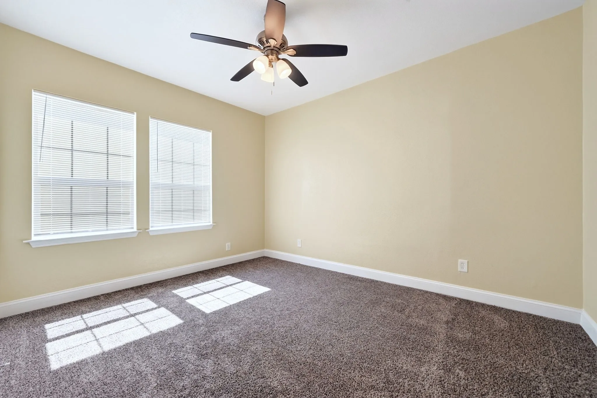 Warm neutral wall paint, two windows with blinds, brown ceiling fan, and carpeted flooring