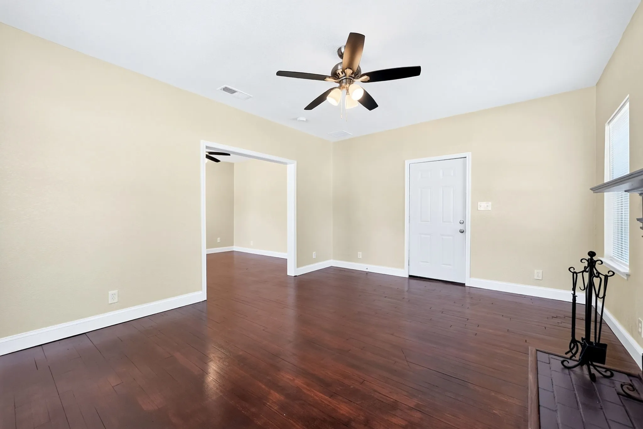 Hardwood flooring, ceiling fan, white paneled door, window with white blinds, and a fireplace with a mantel