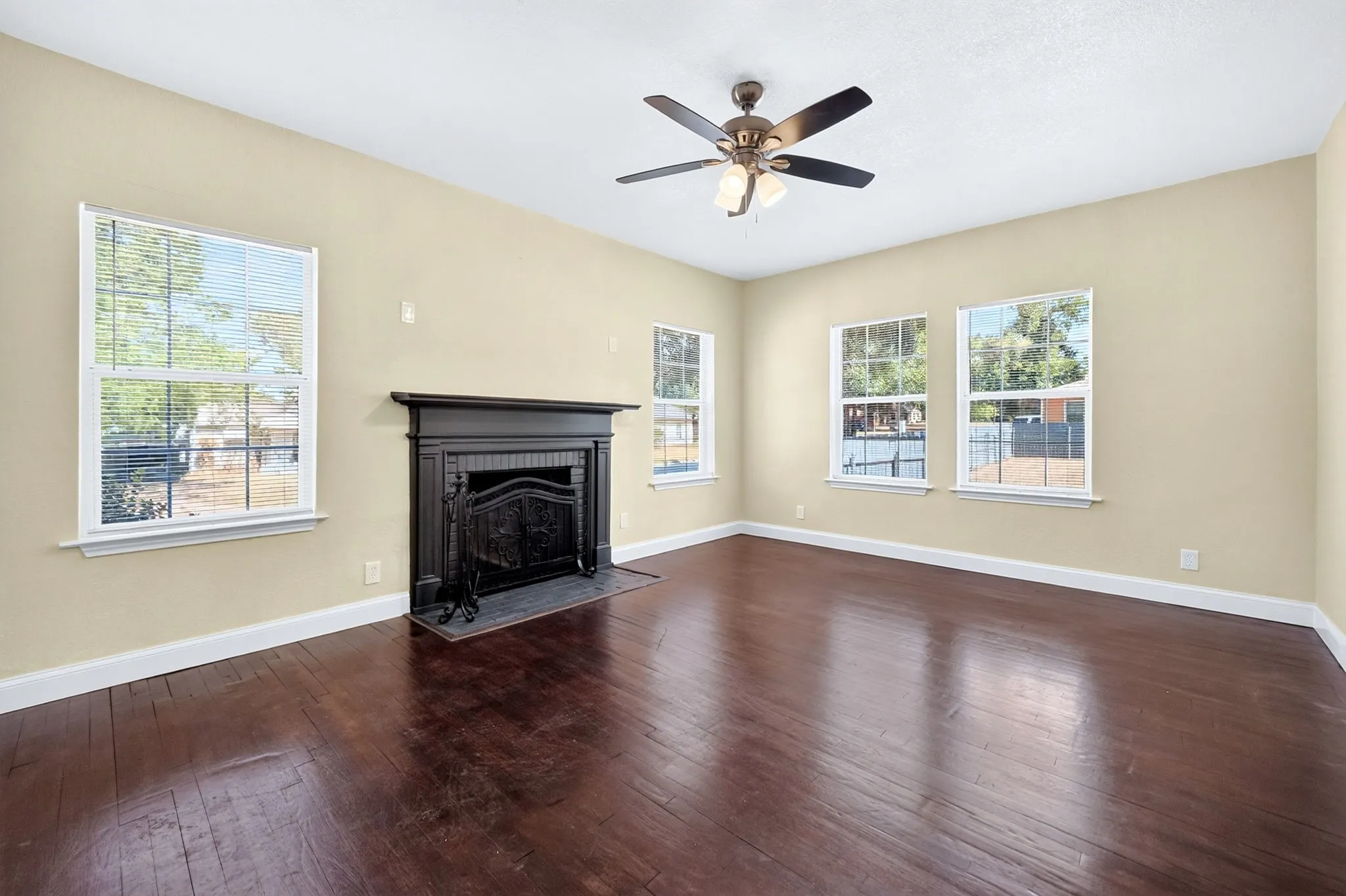 Spacious room featuring rich wood-finish flooring, a traditional fireplace with a decorative mantel, multiple windows, and a ceiling fan with integrated lighting