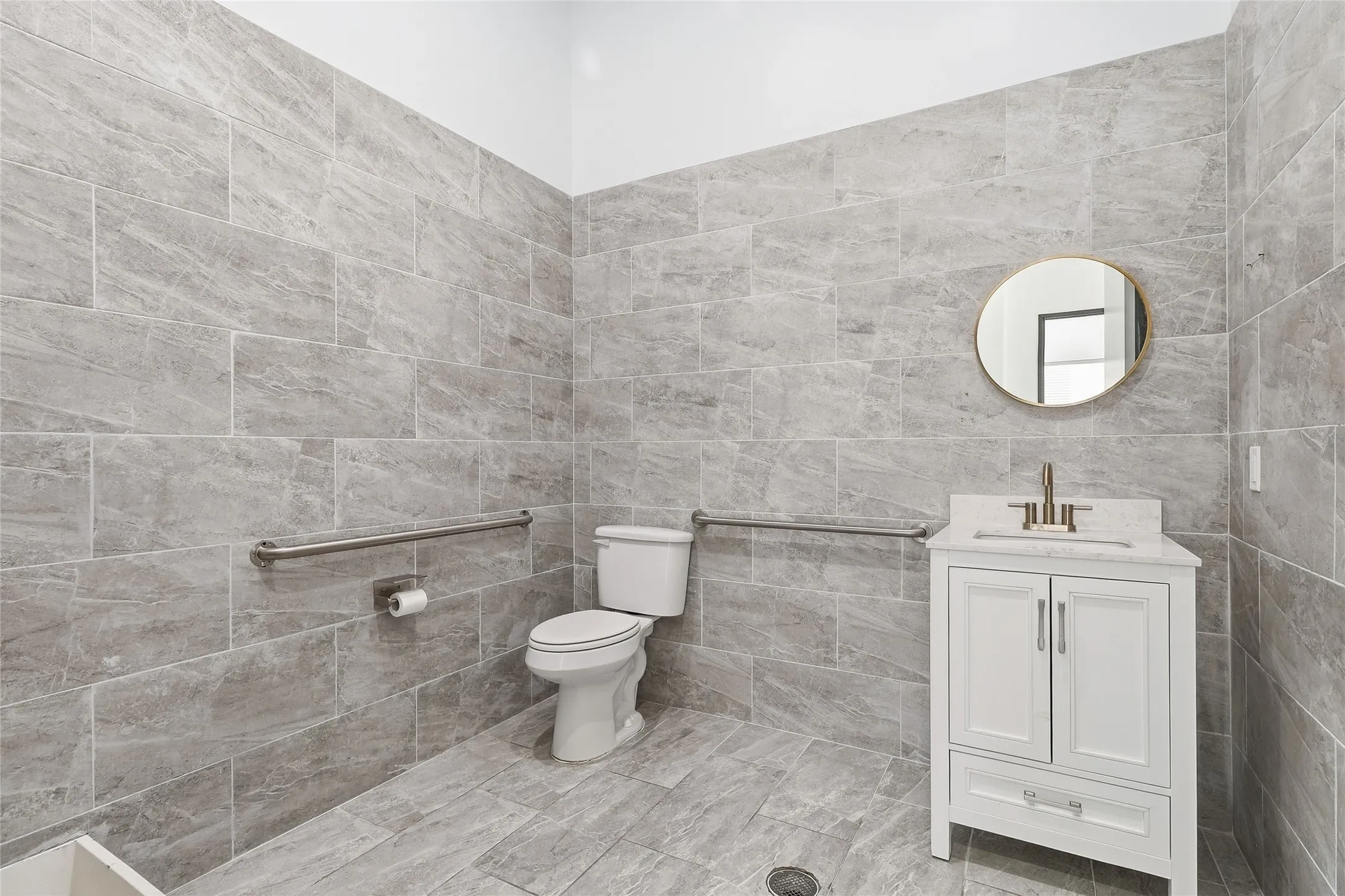 Full wall and floor tile, vanity with marble-finish countertop, round mirror, and grab bars