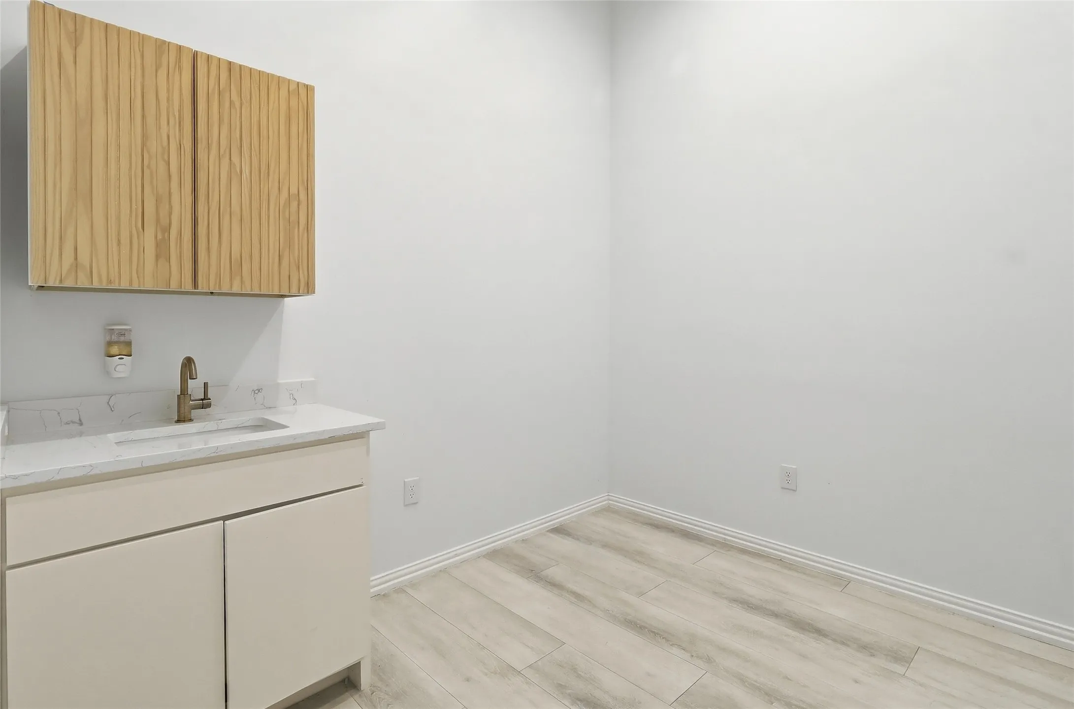 White walls with wood-finish flooring, featuring a built-in vanity with a single basin sink, a wall-mounted faucet, and upper wood-finish cabinetry
