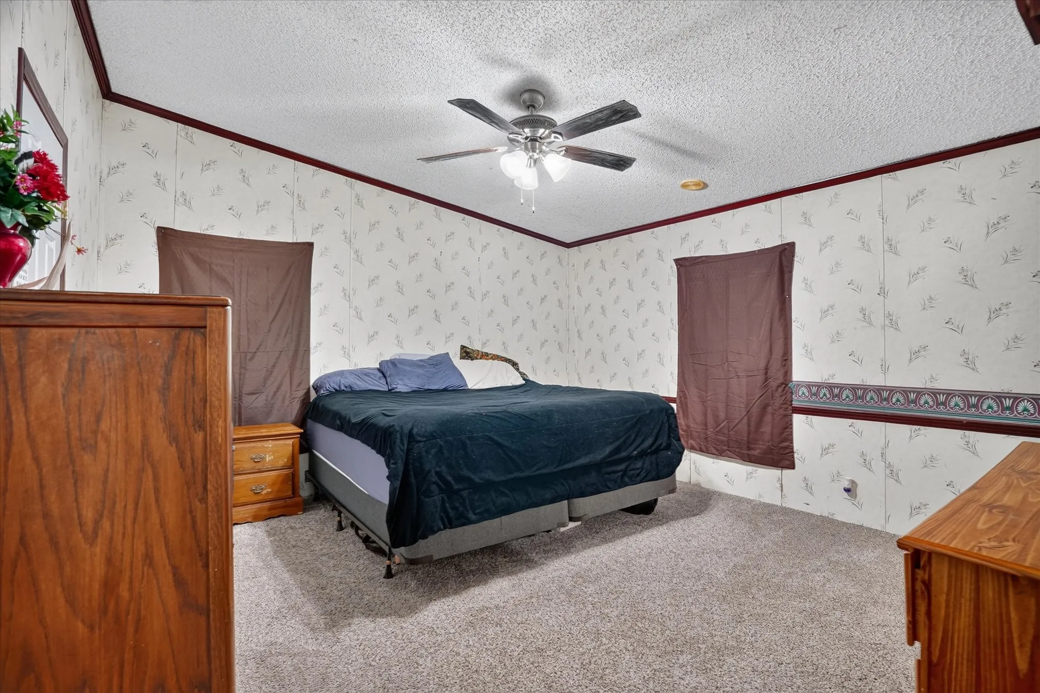 Spacious primary bedroom featuring carpeted flooring, decorative wallpapered walls with coordinating border trim, ceiling fan with integrated lighting, and wood-finish bedroom furnishings