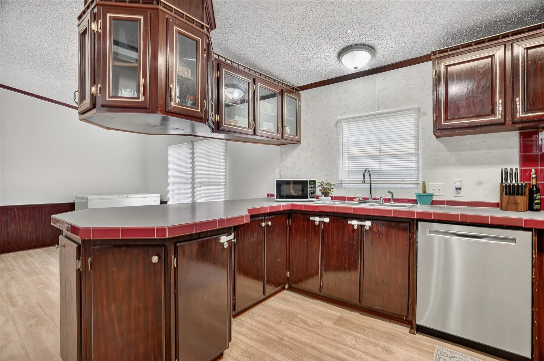 Kitchen featuring wood-finish flooring, dark wood cabinetry with gold-tone hardware, a stainless steel dishwasher, a built-in microwave, and a red tiled backsplash