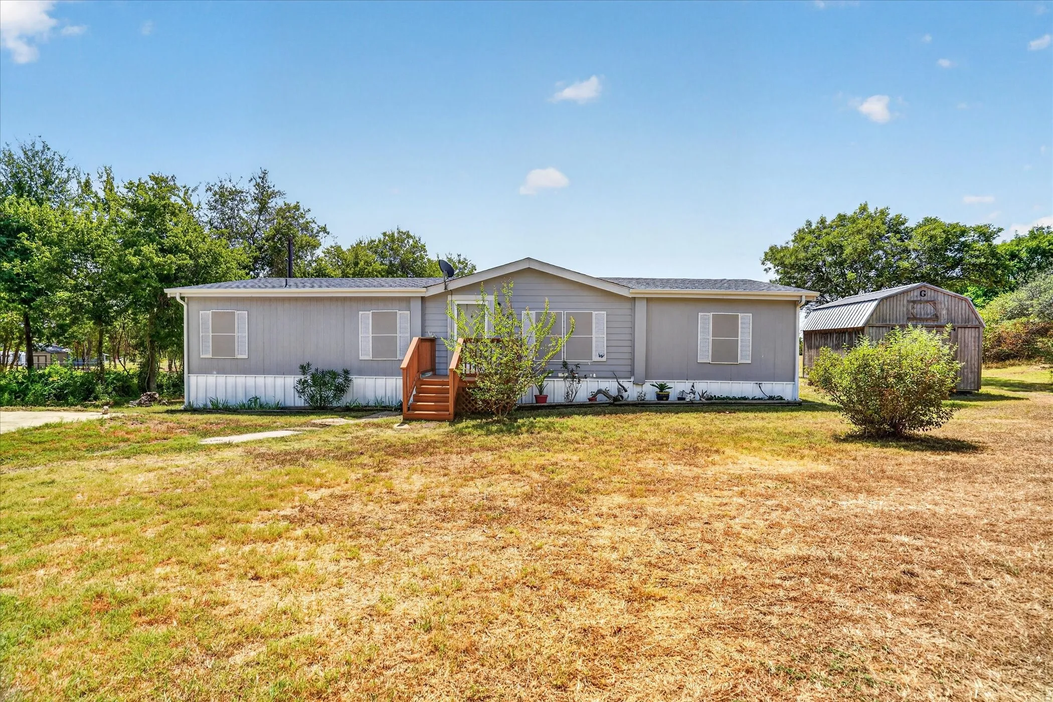 Gray siding exterior with white window shutters, wood-finish porch steps, and a detached outbuilding