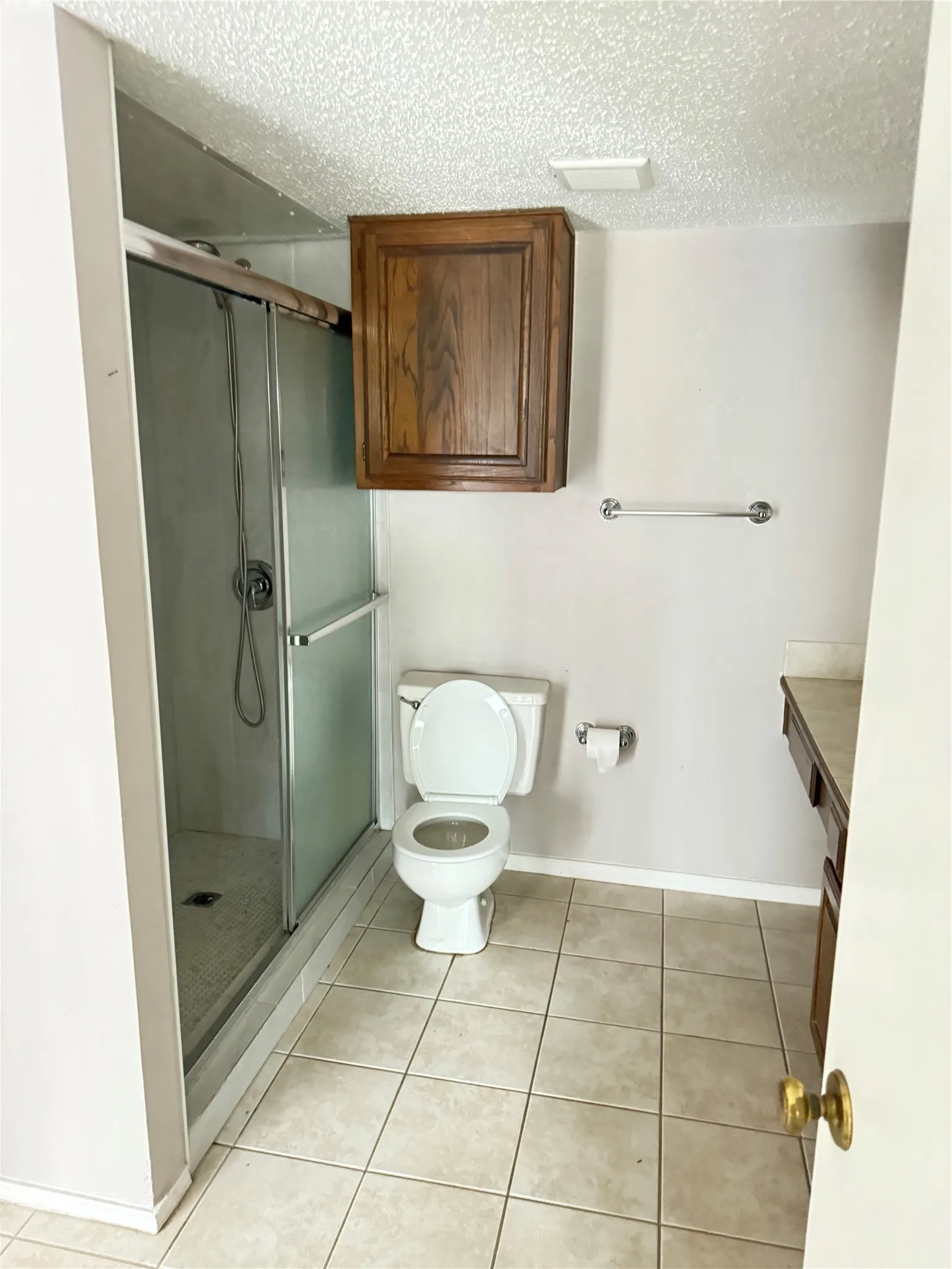 Glass-enclosed shower with a tiled floor, white toilet, single vanity with wood-finish cabinetry, and neutral tile flooring