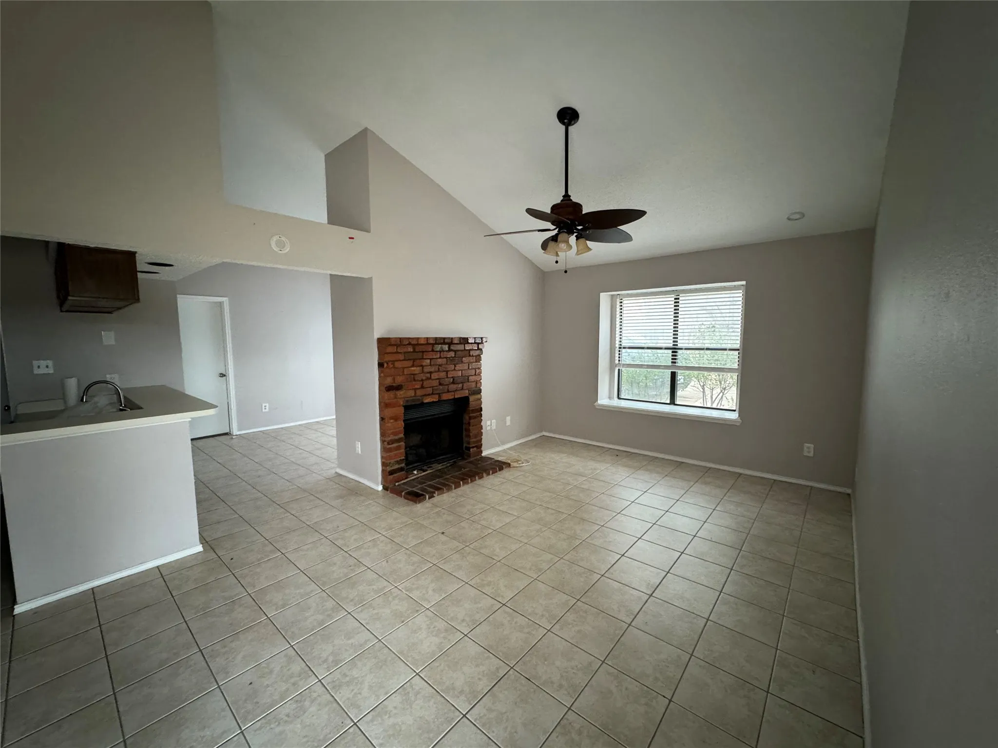 Vaulted ceiling, brick fireplace, tile flooring, window with blinds, and a ceiling fan