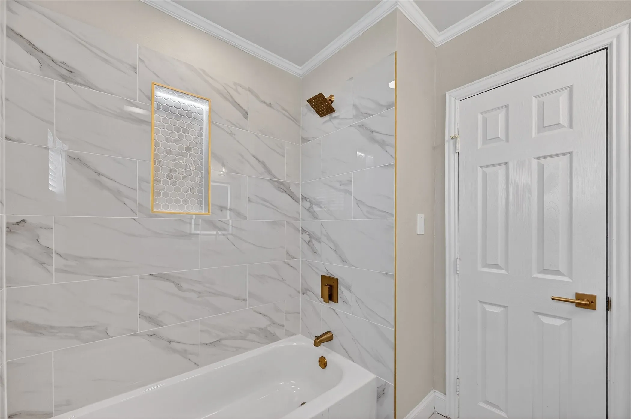 Bathtub with a built-in shower, large format wall tiling, a tiled shower niche with accent mosaic, and gold-finish fixtures
