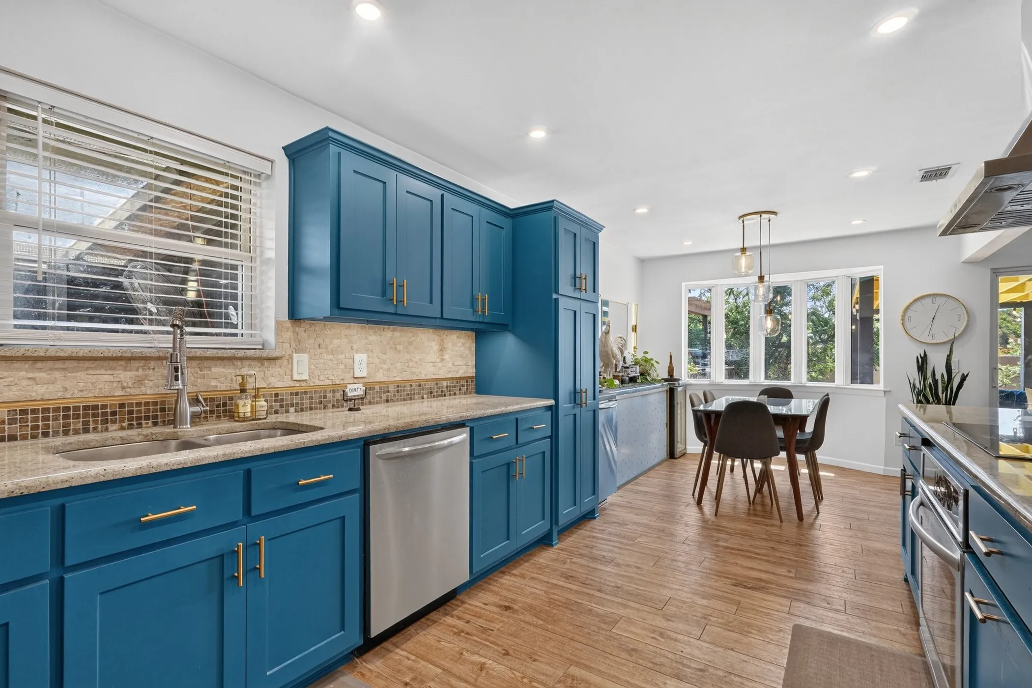 Blue cabinetry with gold-tone hardware, stainless steel appliances, stone countertops, mosaic tile backsplash, and wood-finish flooring