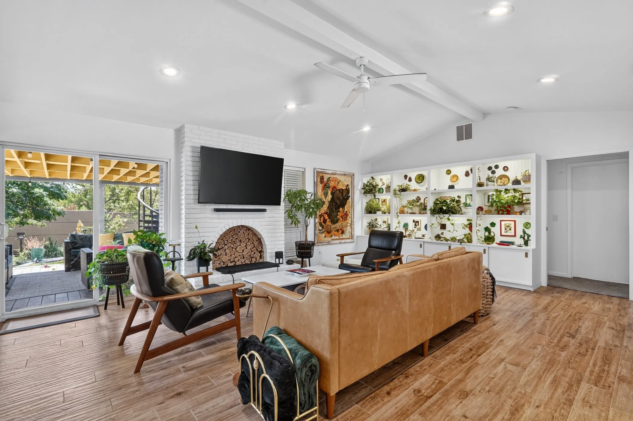 Vaulted ceiling with exposed beams, wood-finish flooring, white brick fireplace, built-in illuminated shelving, and sliding glass door to exterior