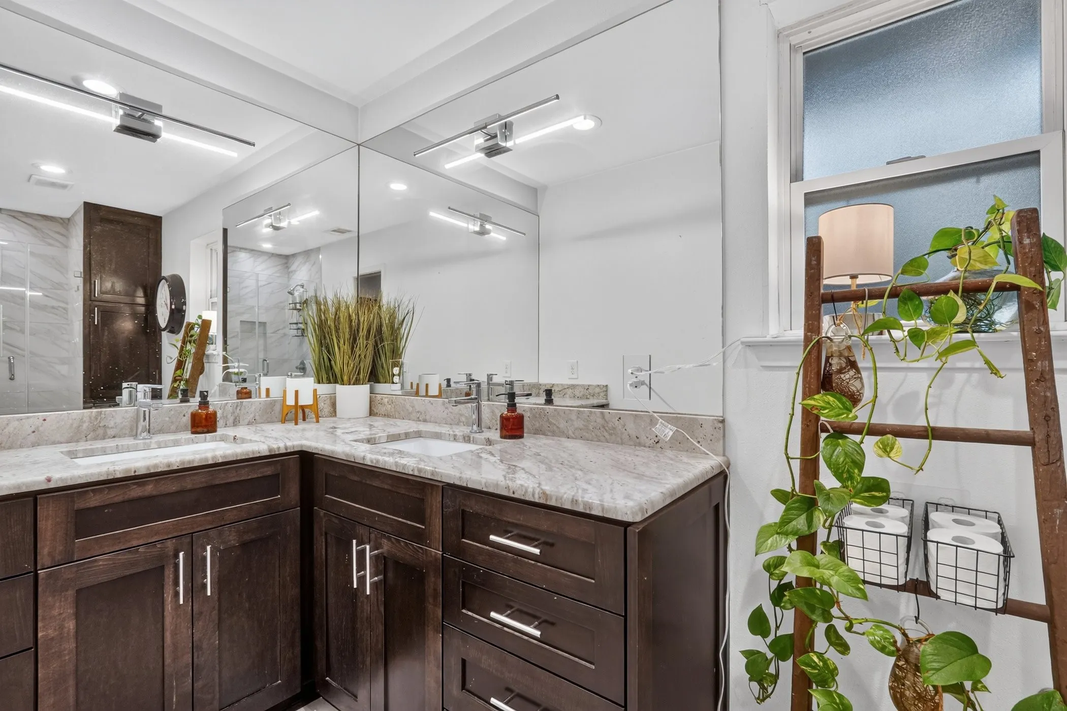 Corner vanity with dual integrated sinks and light-toned countertop, dark wood-finish cabinetry, full-wall mirror, and wall-mounted lighting fixtures