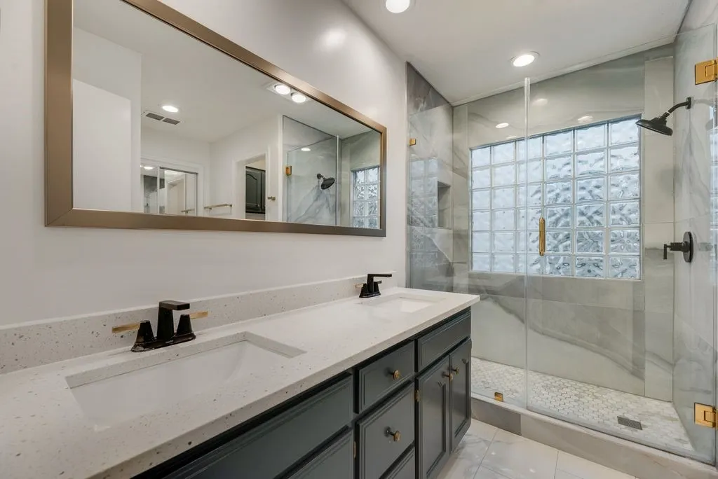Dual vanity with composite countertop, frameless glass shower enclosure, glass block window, and tile shower surround
