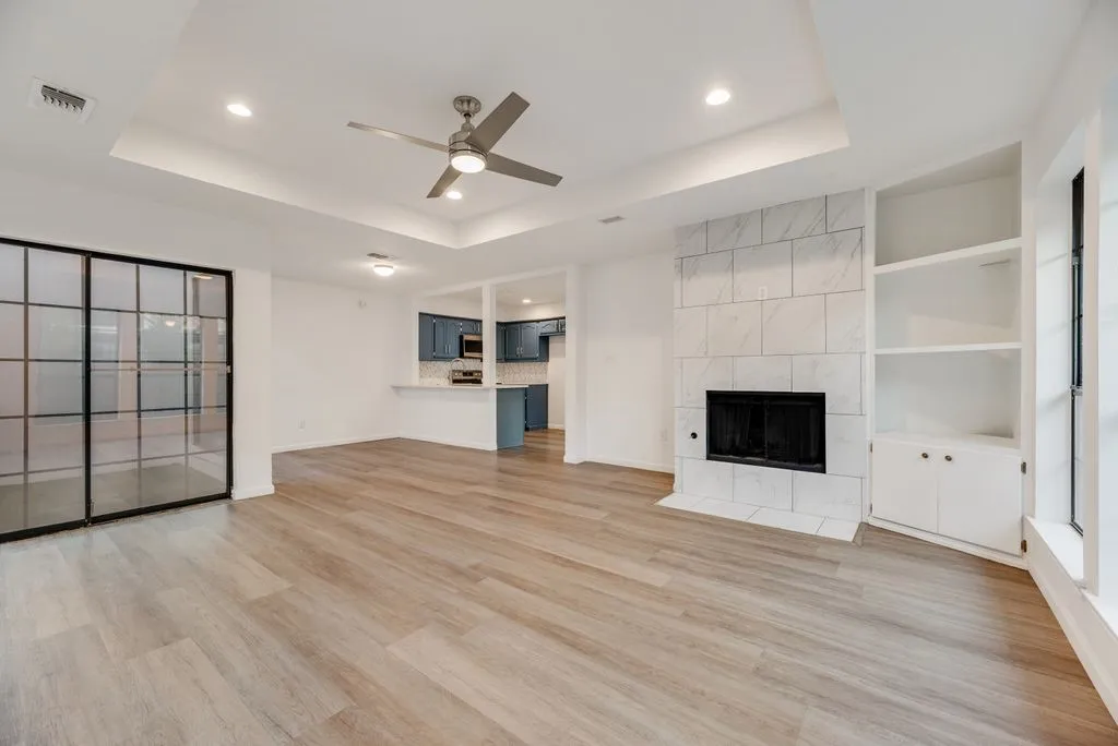 Wood-finish flooring, recessed lighting, tray ceiling with ceiling fan, tiled fireplace, and built-in shelving with cabinetry