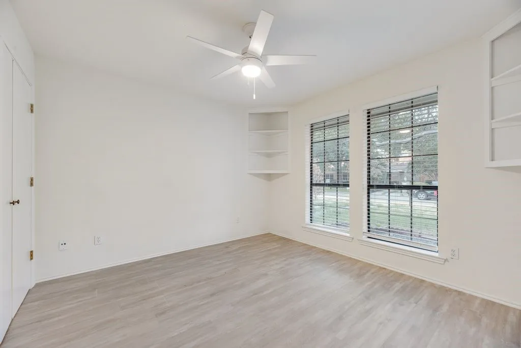 Wood-finish flooring, two large windows, corner built-in shelving, ceiling fan with integrated lighting, and a built-in wall cabinet